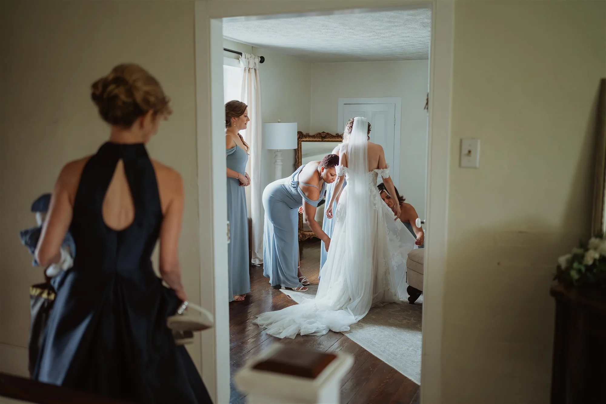 Bridesmaids in blue help bride into wedding gown while guest looks on through doorway