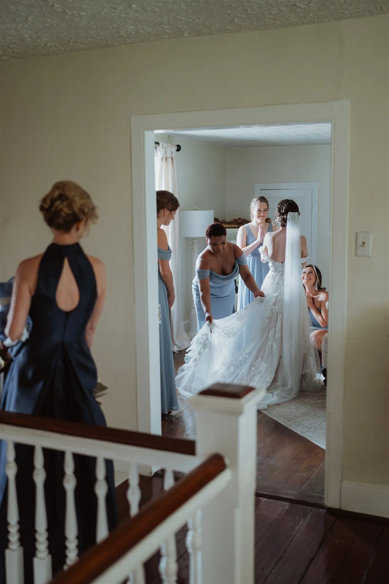 Bridesmaids in blue help bride into her gown in a bright upstairs room while guests look on from the hallway
