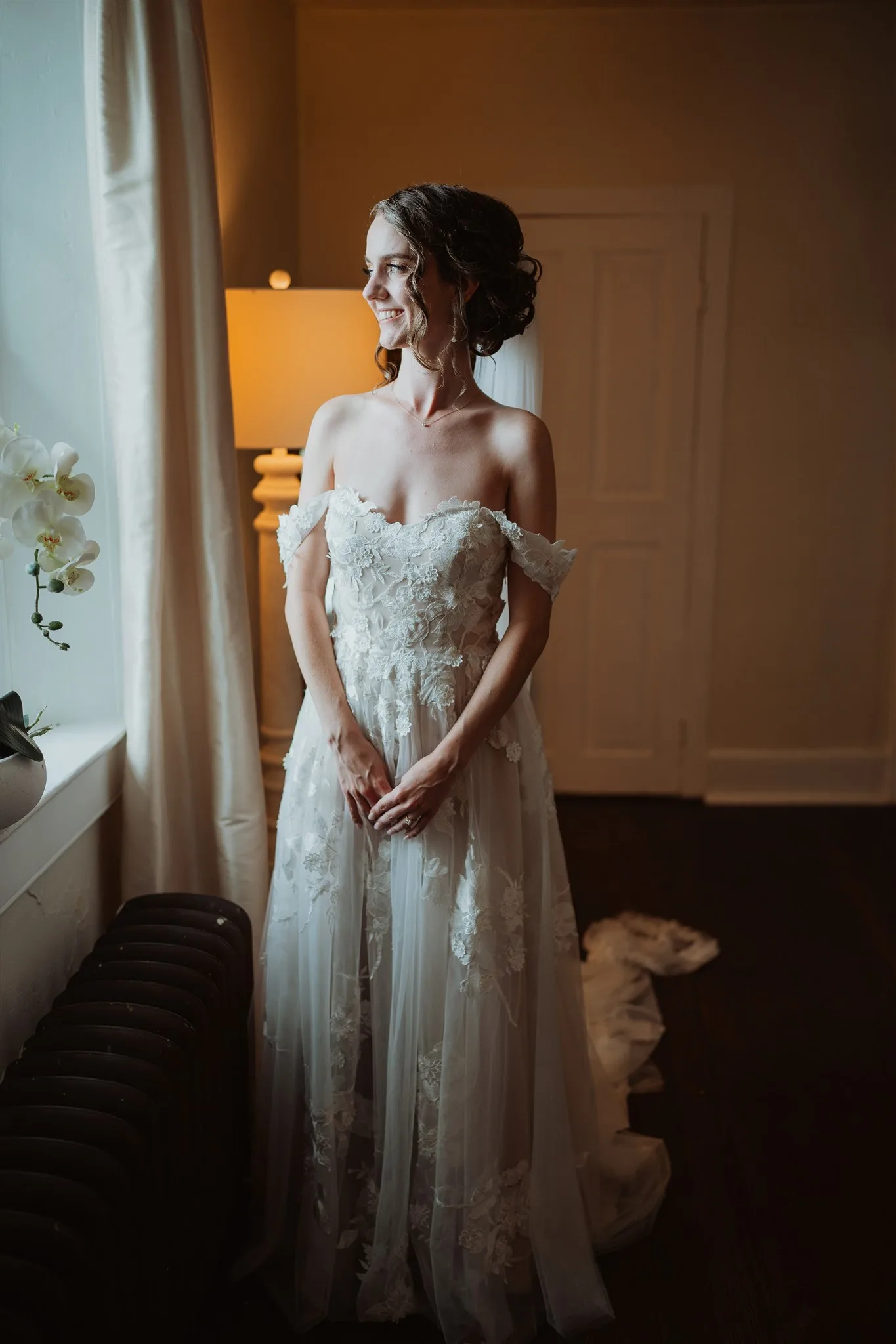 Bride in off-shoulder lace gown smiling by window light in a warmly lit room before her wedding