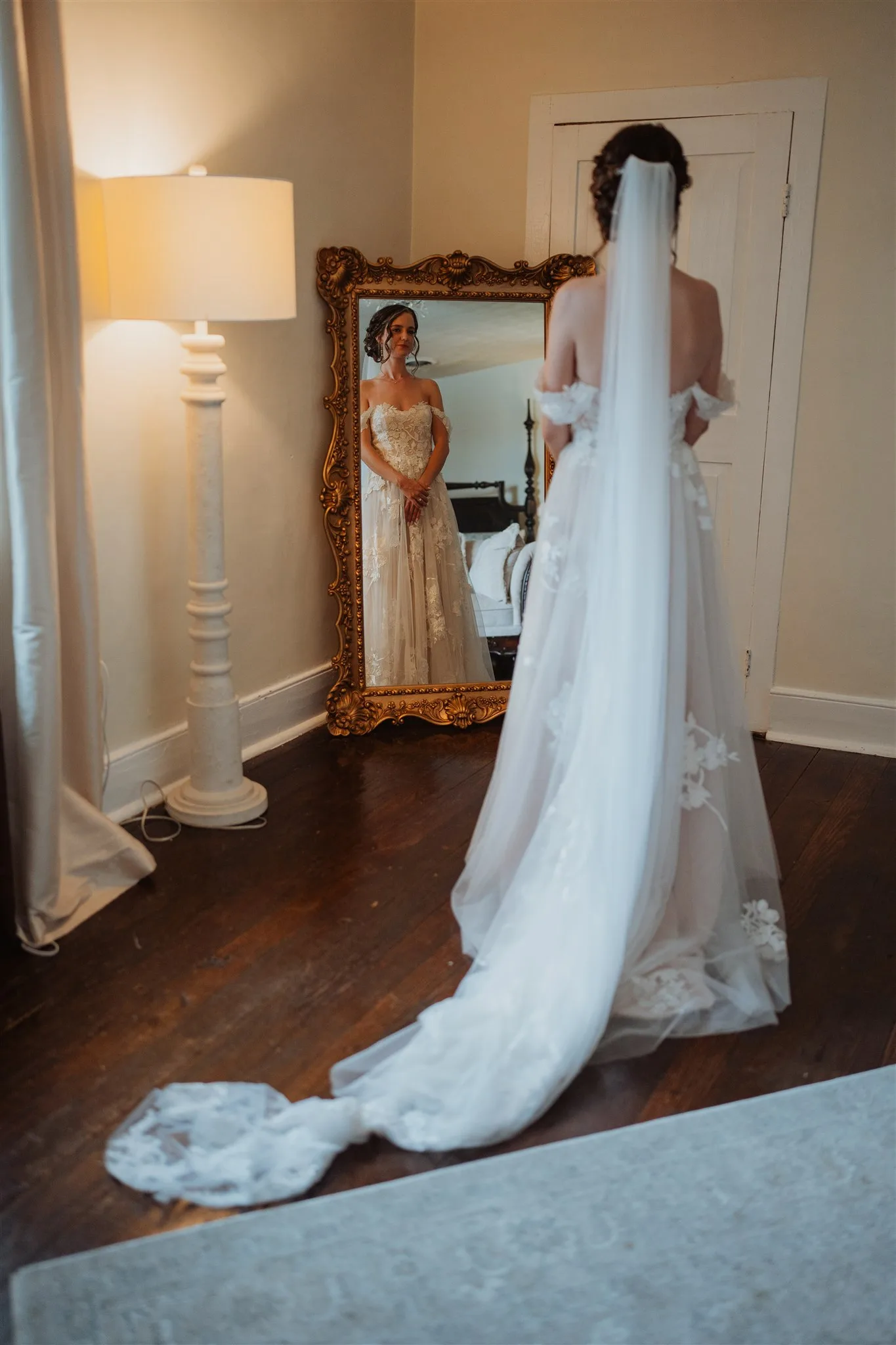 Bride in lace gown and cathedral veil gazes at her reflection in an ornate gold mirror before the ceremony