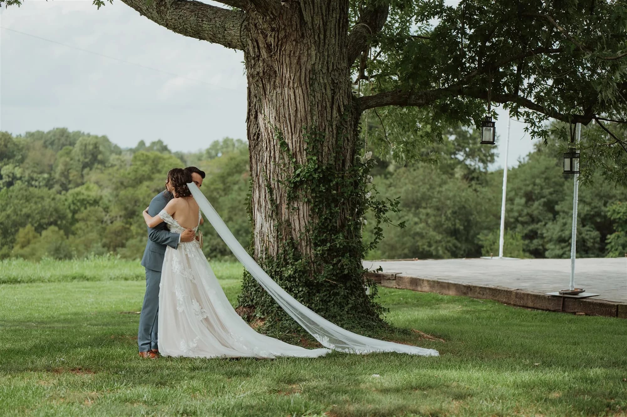 Bride and groom embrace beneath a grand oak tree on Rixey Manor's lush green grounds, cathedral veil trailing across the grass.