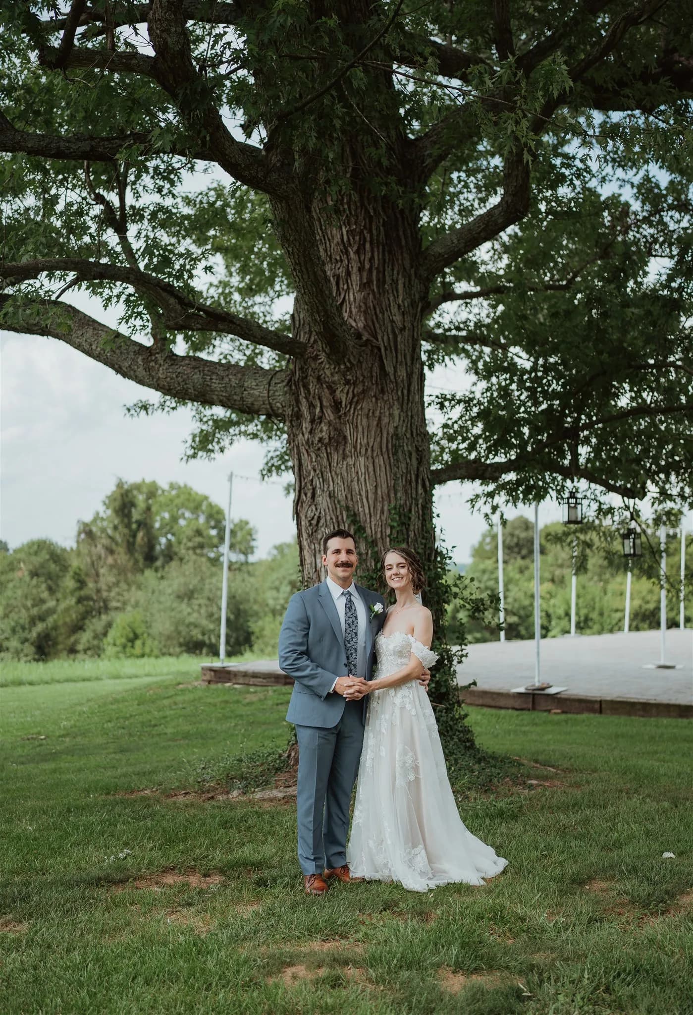 Bride and groom posing hand-in-hand beneath a massive oak tree on the lush Rixey Manor grounds