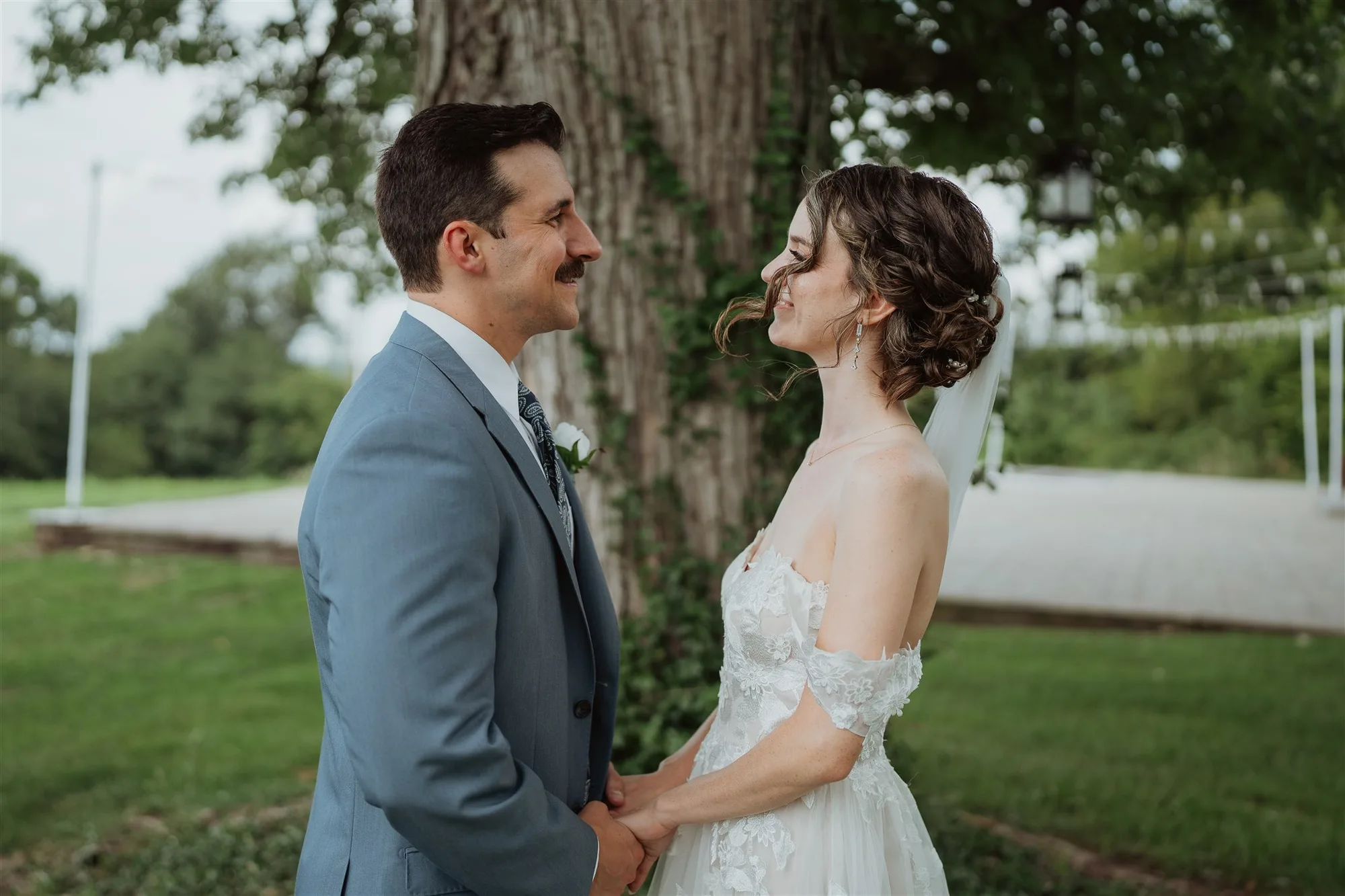 Bride and groom hold hands and share a smile during outdoor portraits beneath a large tree at Rixey Manor