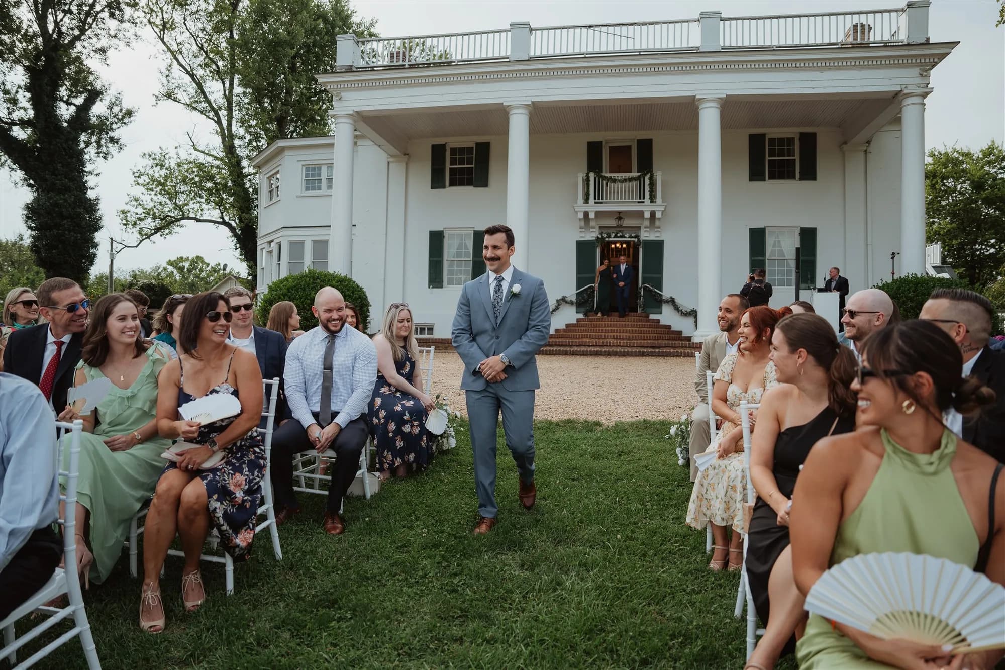 Groom walks the aisle at outdoor ceremony before Rixey Manor's white colonial facade as guests smile