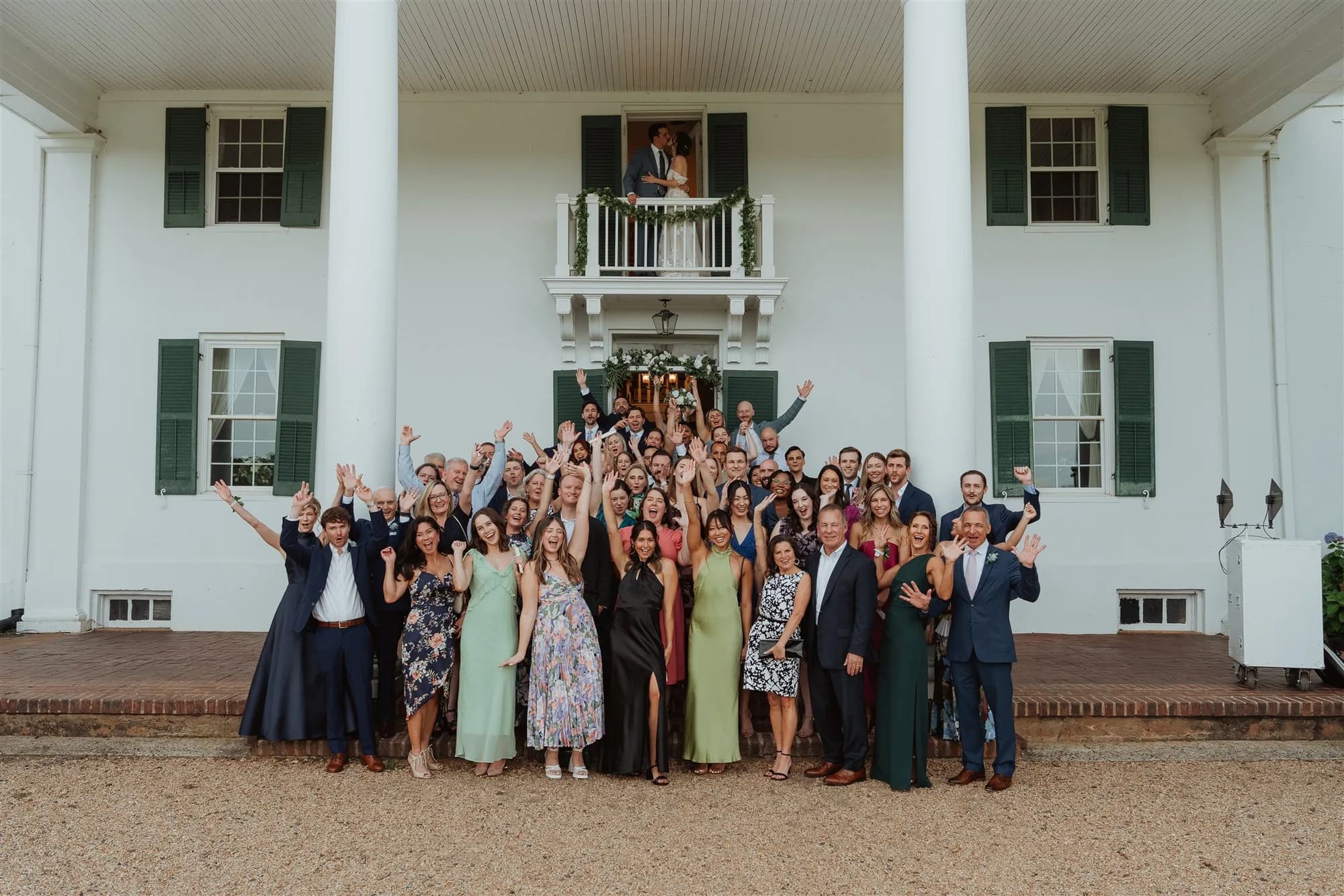 Large wedding party group photo in front of white colonial manor with green shutters, guests in formal attire celebrating.