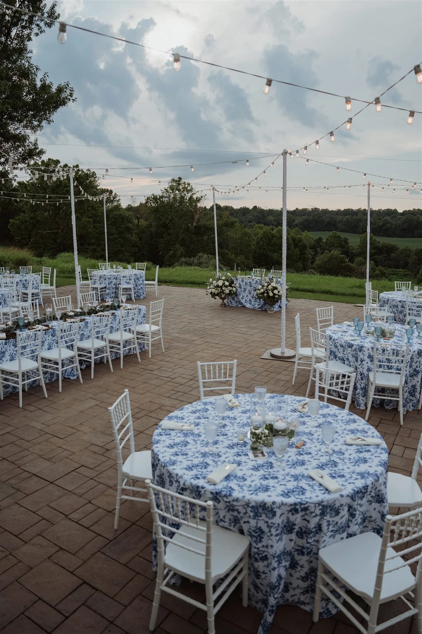 Outdoor wedding reception with blue floral tablecloths, white chiavari chairs, and string lights at rural venue