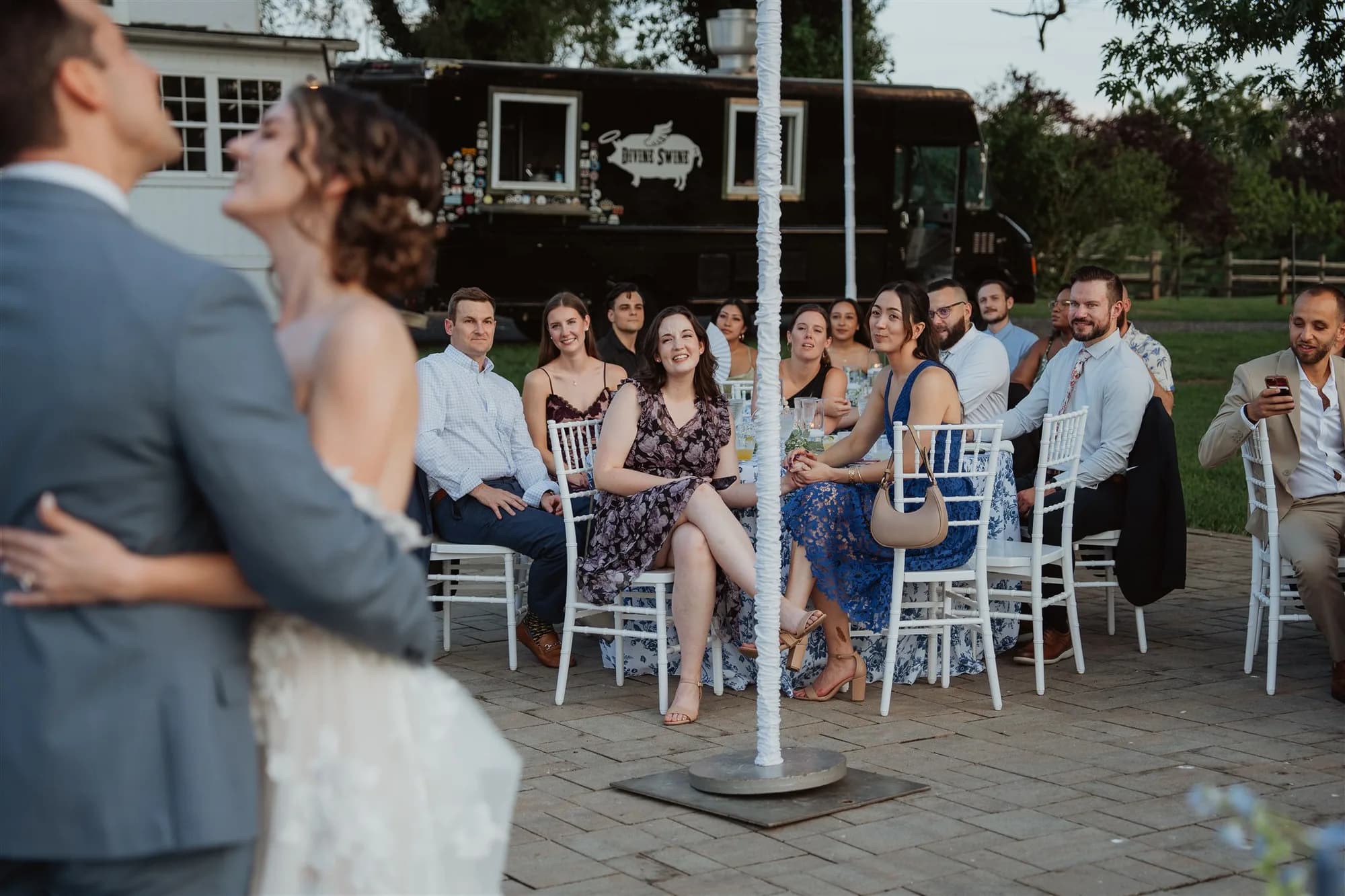 Couple shares first dance outdoors as smiling guests watch from white chairs at a summer wedding reception