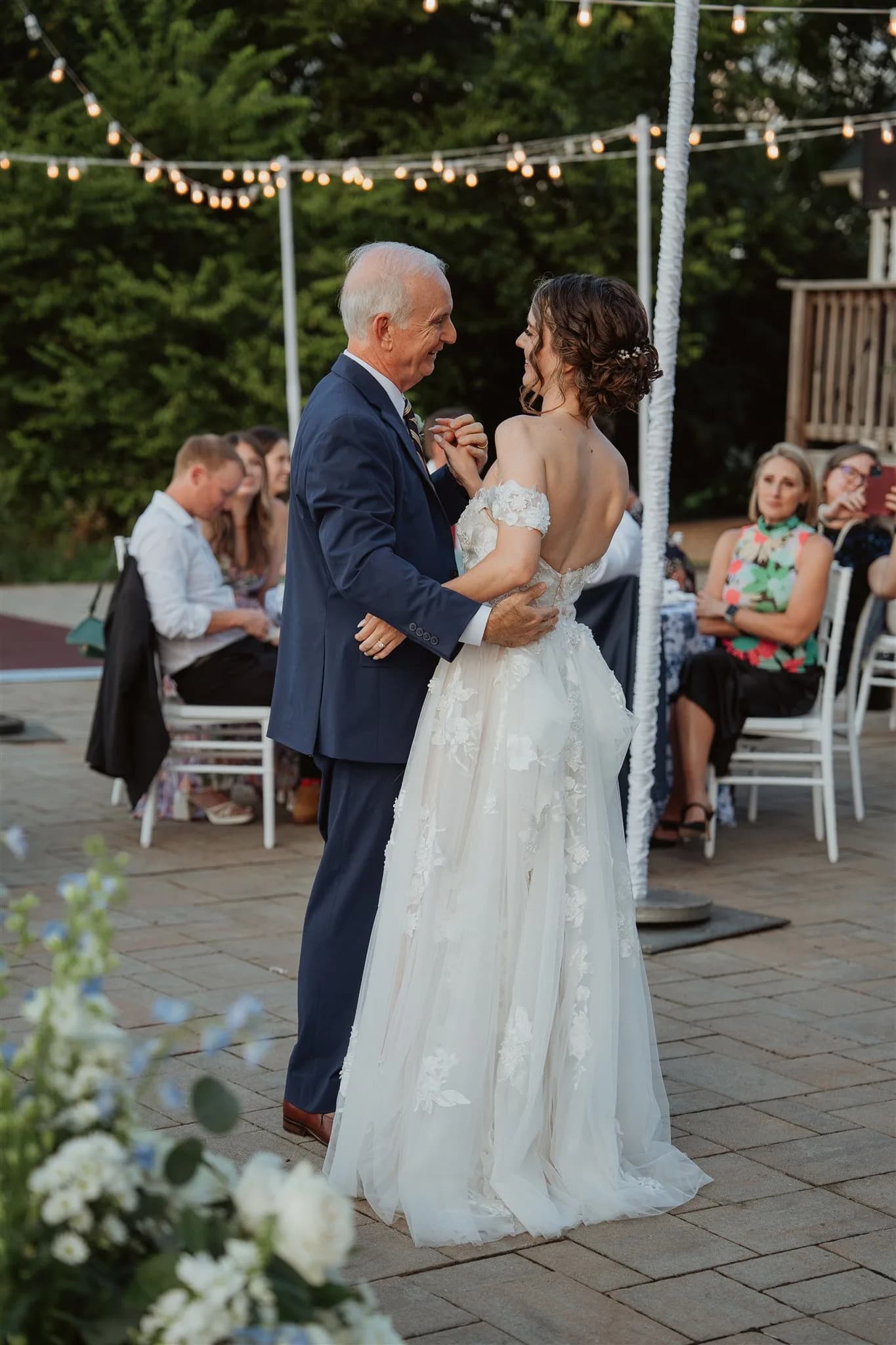 Bride shares father-daughter dance on outdoor patio under string lights at dusk wedding reception