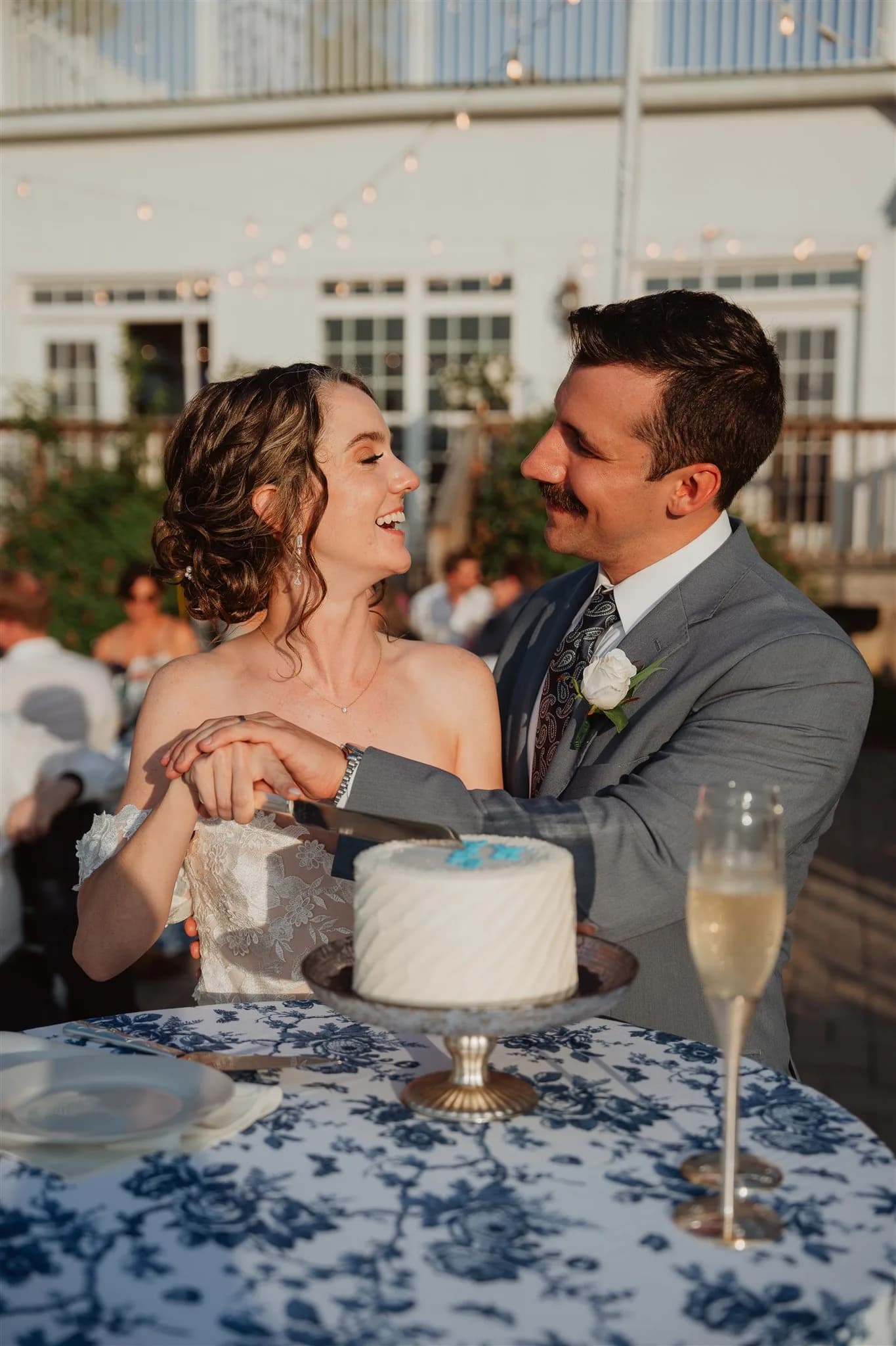 Bride and groom laugh together while cutting their white wedding cake on the Rixey Manor terrace at golden hour