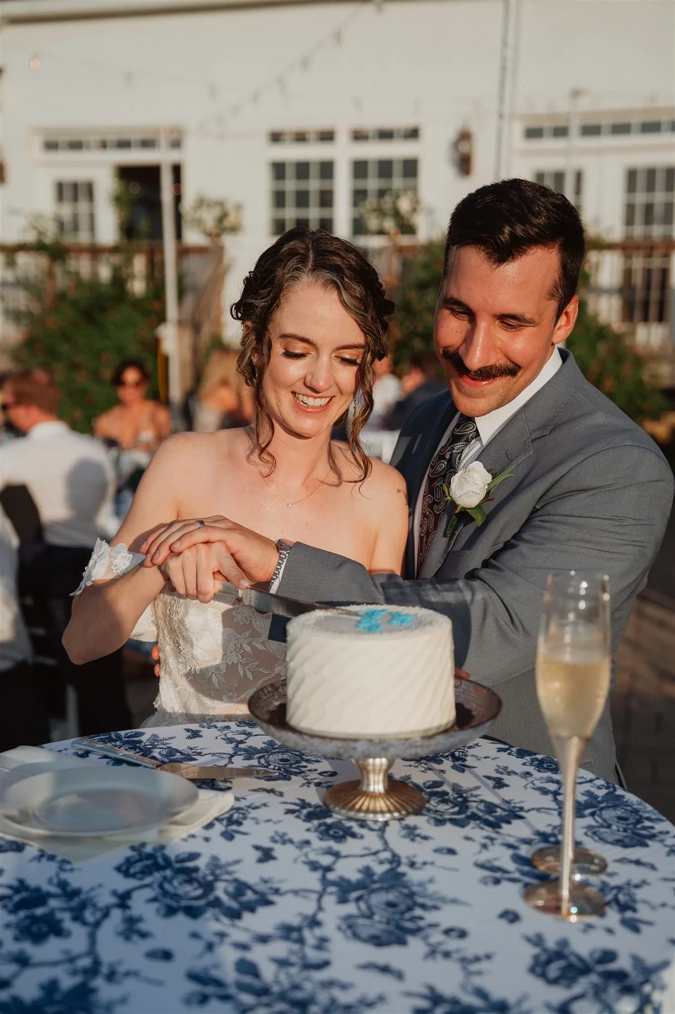 Smiling bride and groom cut their white wedding cake together at an outdoor reception table