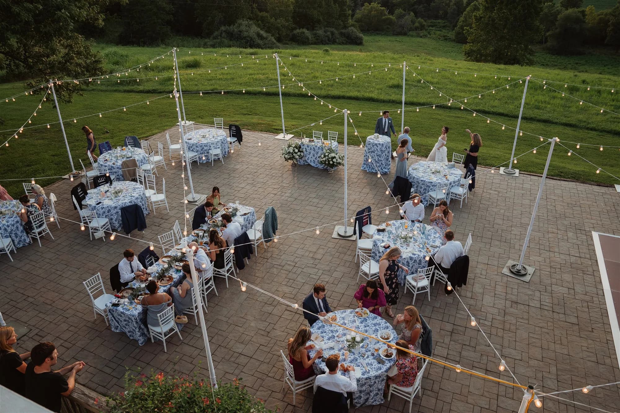 Aerial view of outdoor reception on Rixey Manor terrace with string lights, blue floral linens, and lush green grounds