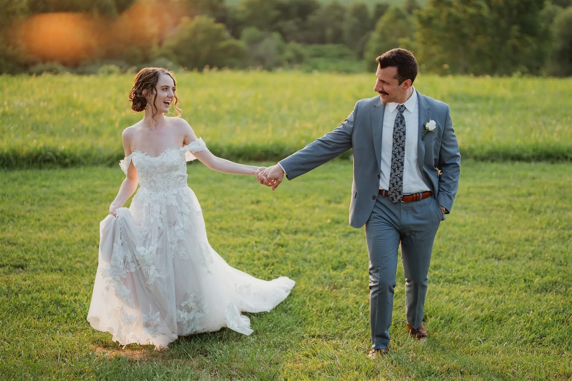 Bride and groom holding hands in green field, bride in white off-shoulder gown, groom in blue suit