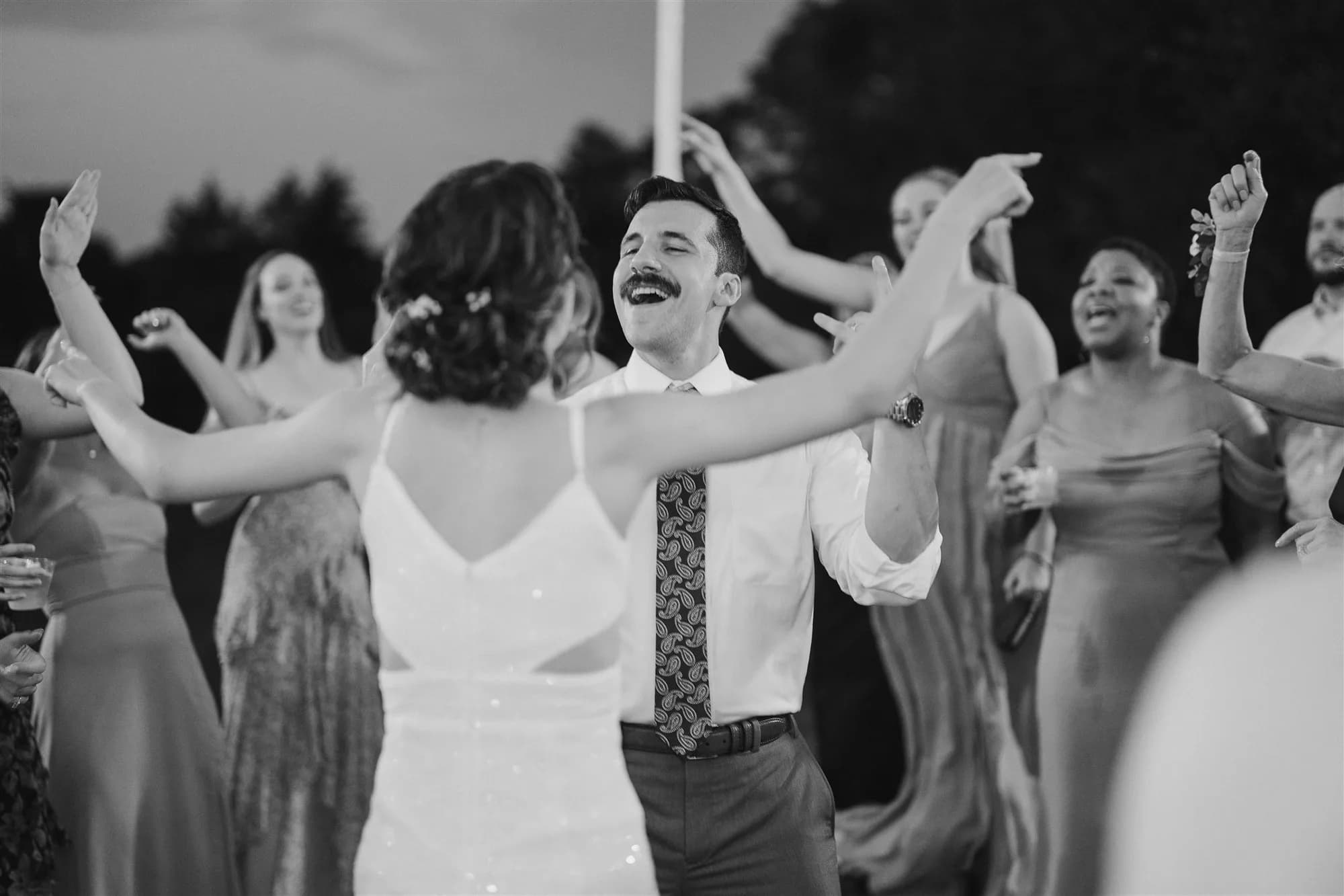 Groom laughs joyfully while dancing with bride surrounded by celebrating guests at outdoor wedding reception
