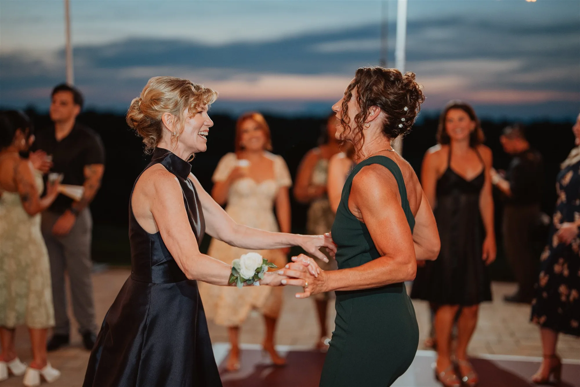 Two women laugh and dance together at an outdoor wedding reception at twilight