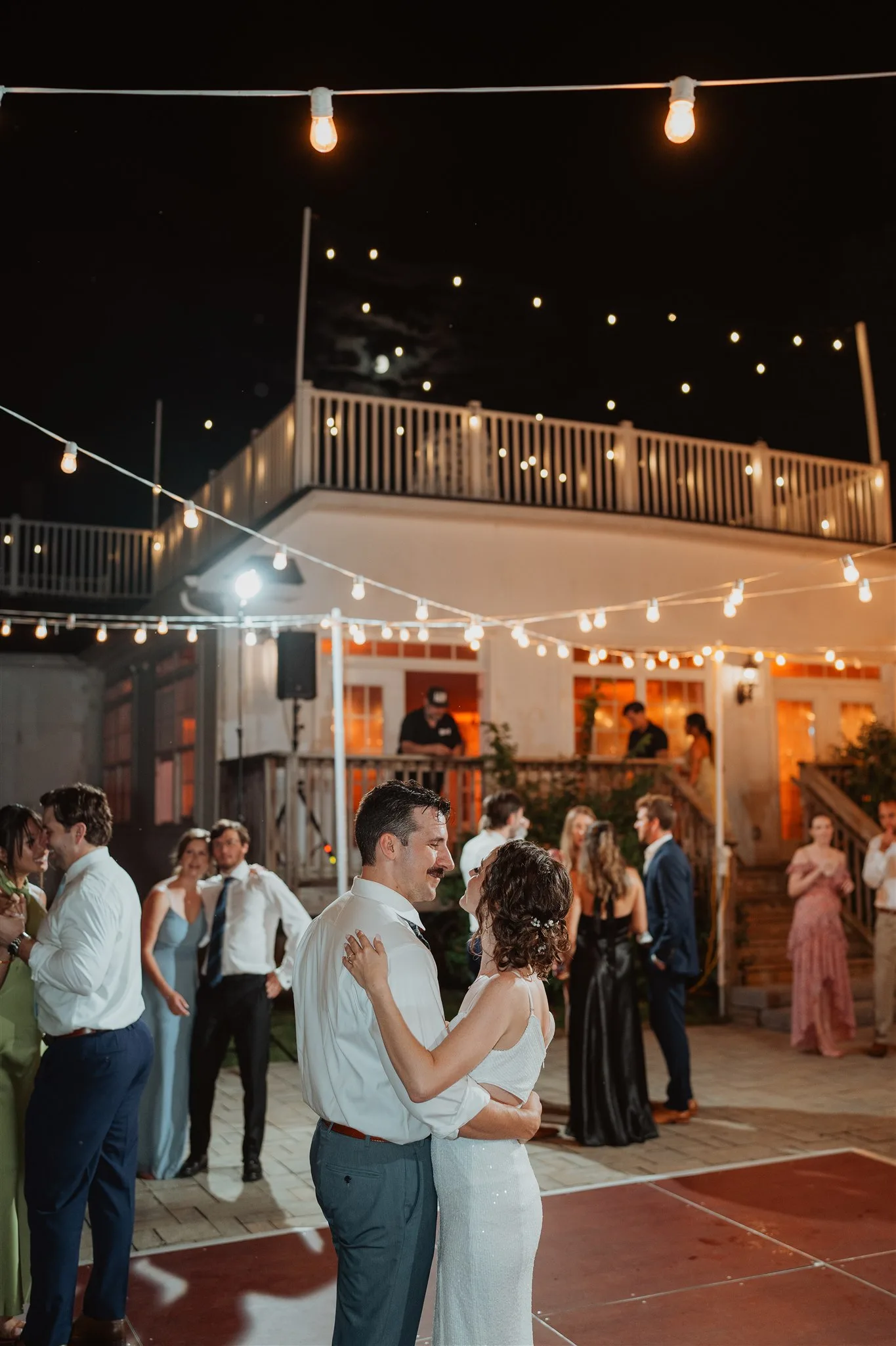 Couple shares first dance under string lights on outdoor patio at night, guests watching around them