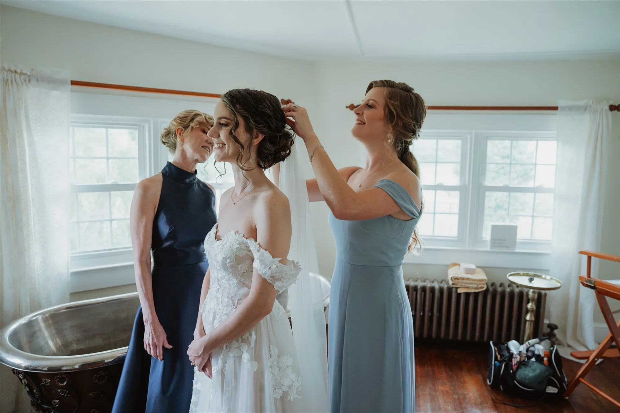 Bride in white dress with two bridesmaids adjusting her hair in a bright getting-ready room