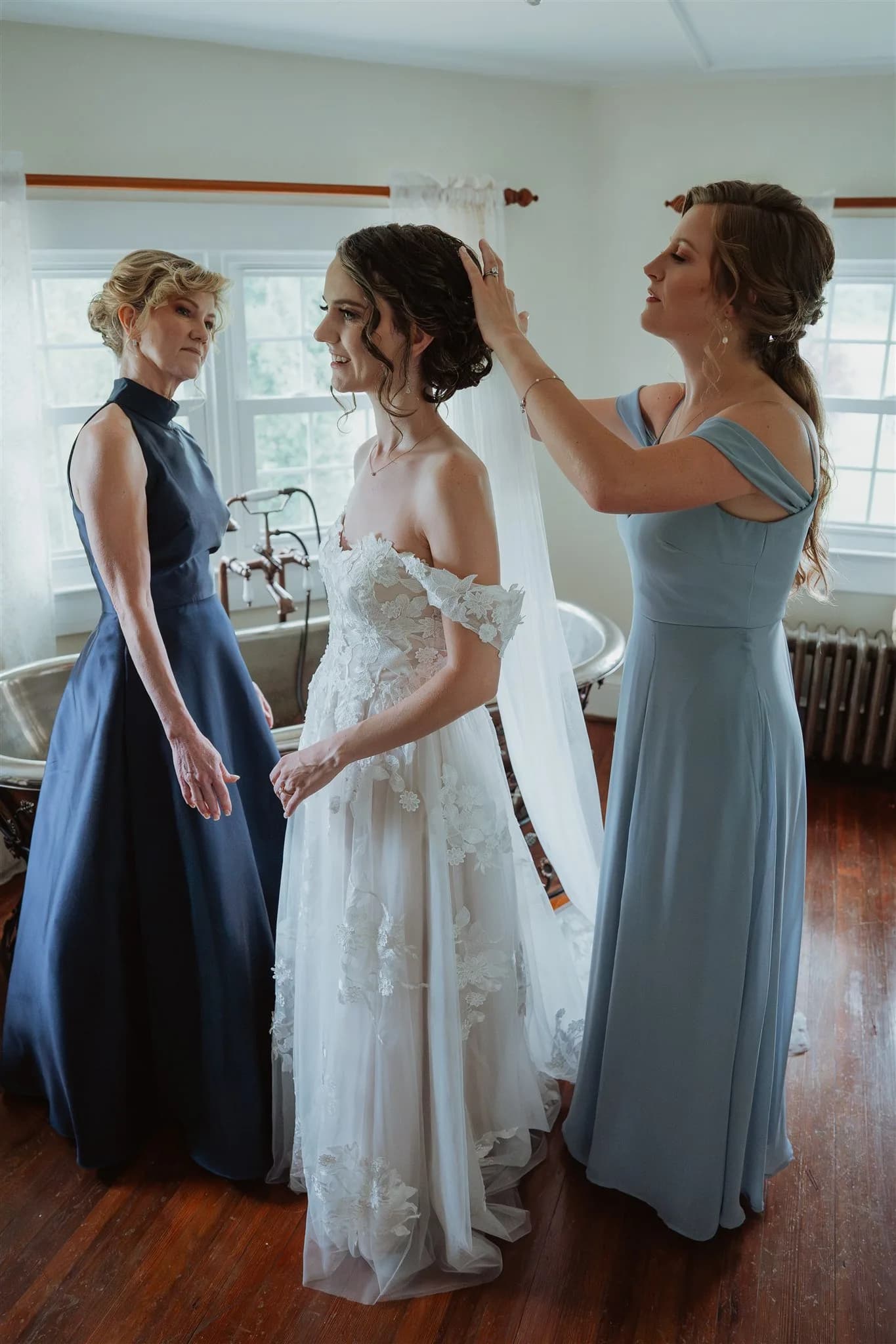 Bride smiles as bridesmaids help place her veil in a sunlit getting-ready room at Rixey Manor