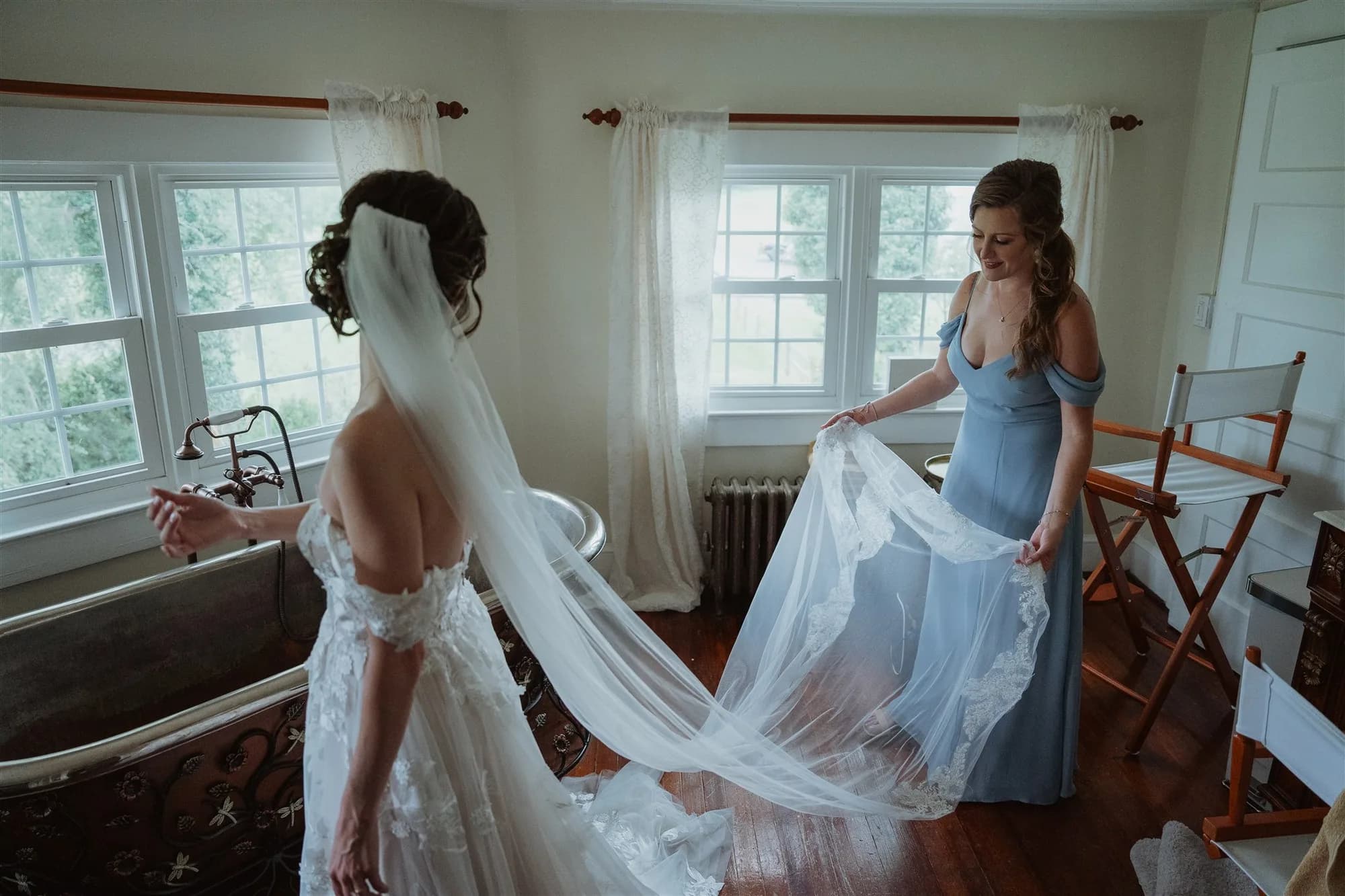 Bridesmaid in blue gown arranges bride's lace veil and train in a sunlit room at Rixey Manor
