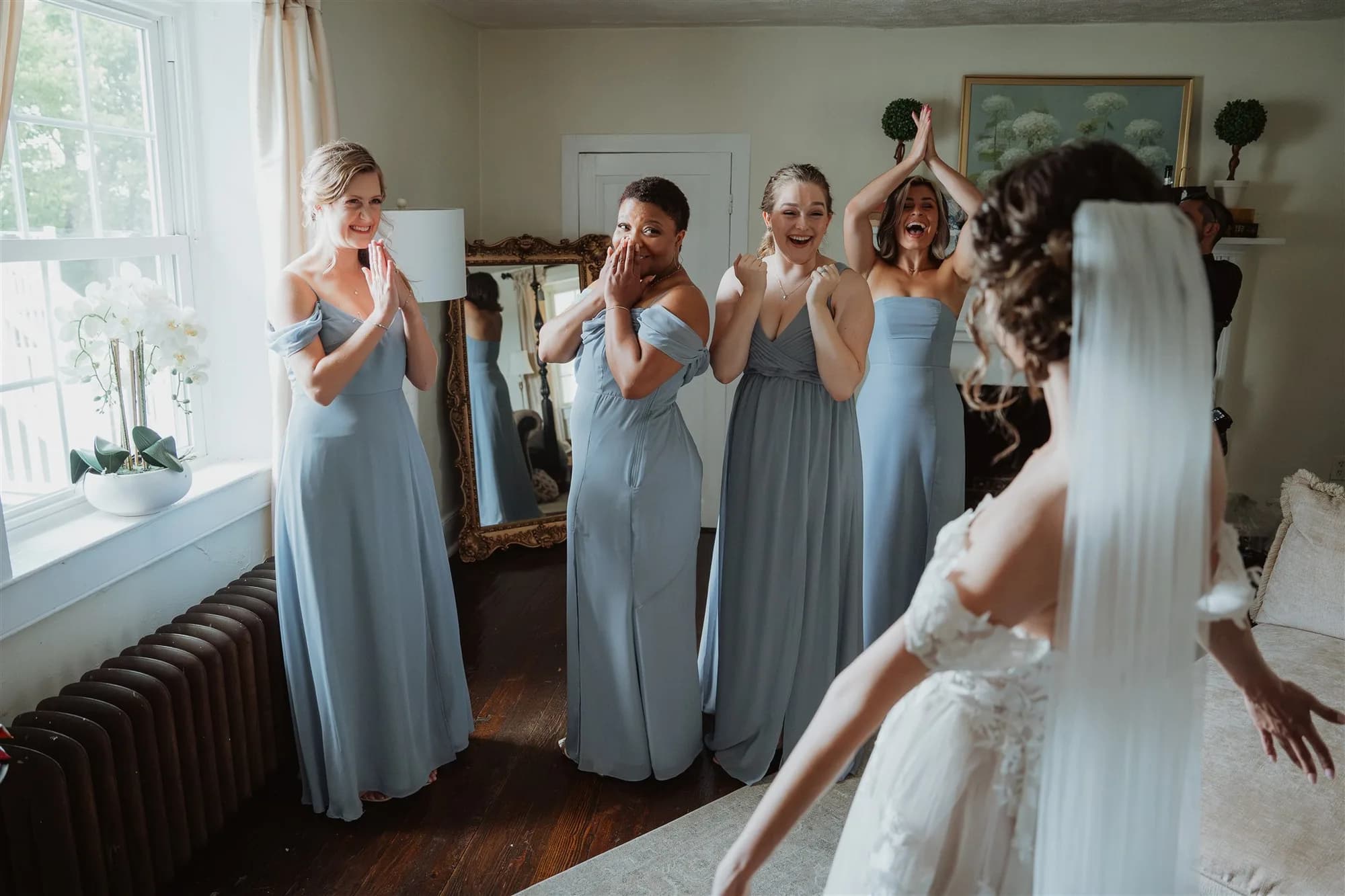 Bridesmaids in blue dresses react with joy and tears seeing the bride in her wedding gown for the first time