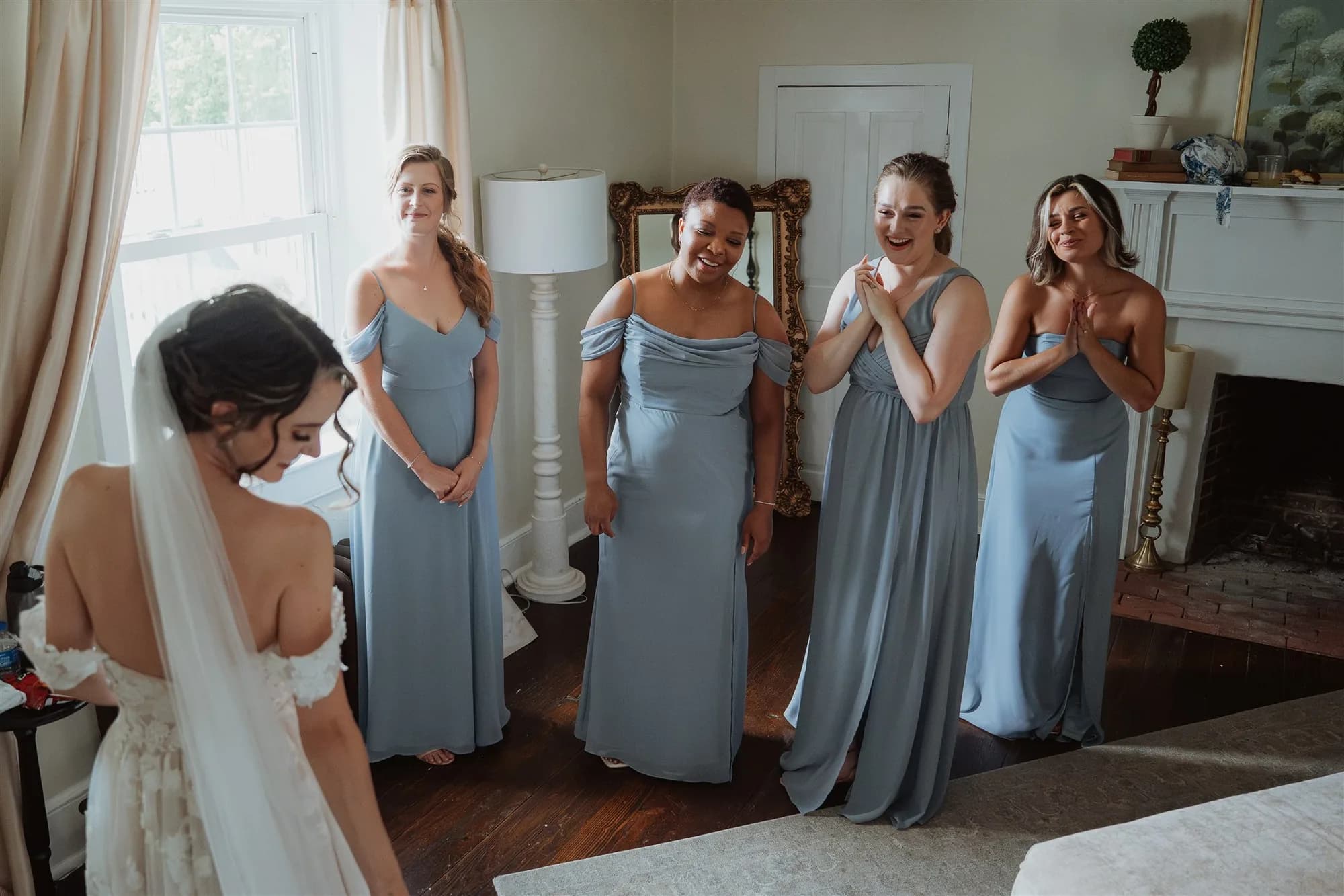 Bridesmaids in dusty blue gowns react with joy seeing bride in wedding dress during getting-ready moment