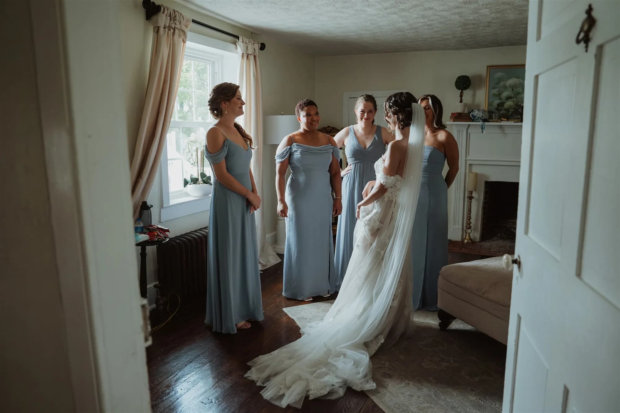 Bride in white gown with long veil stands with four bridesmaids in blue dresses in a manor bedroom