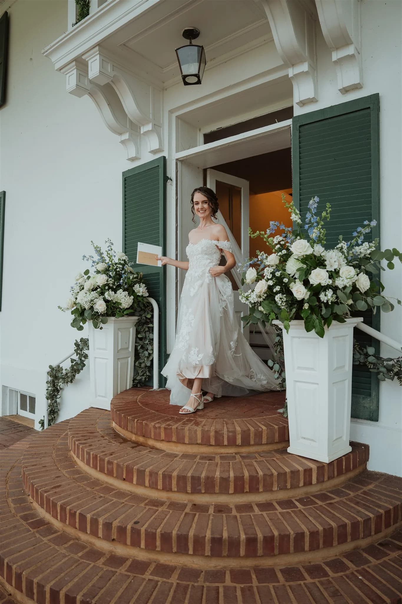 Smiling bride in off-shoulder gown descends Rixey Manor's brick carriage steps flanked by white floral arrangements