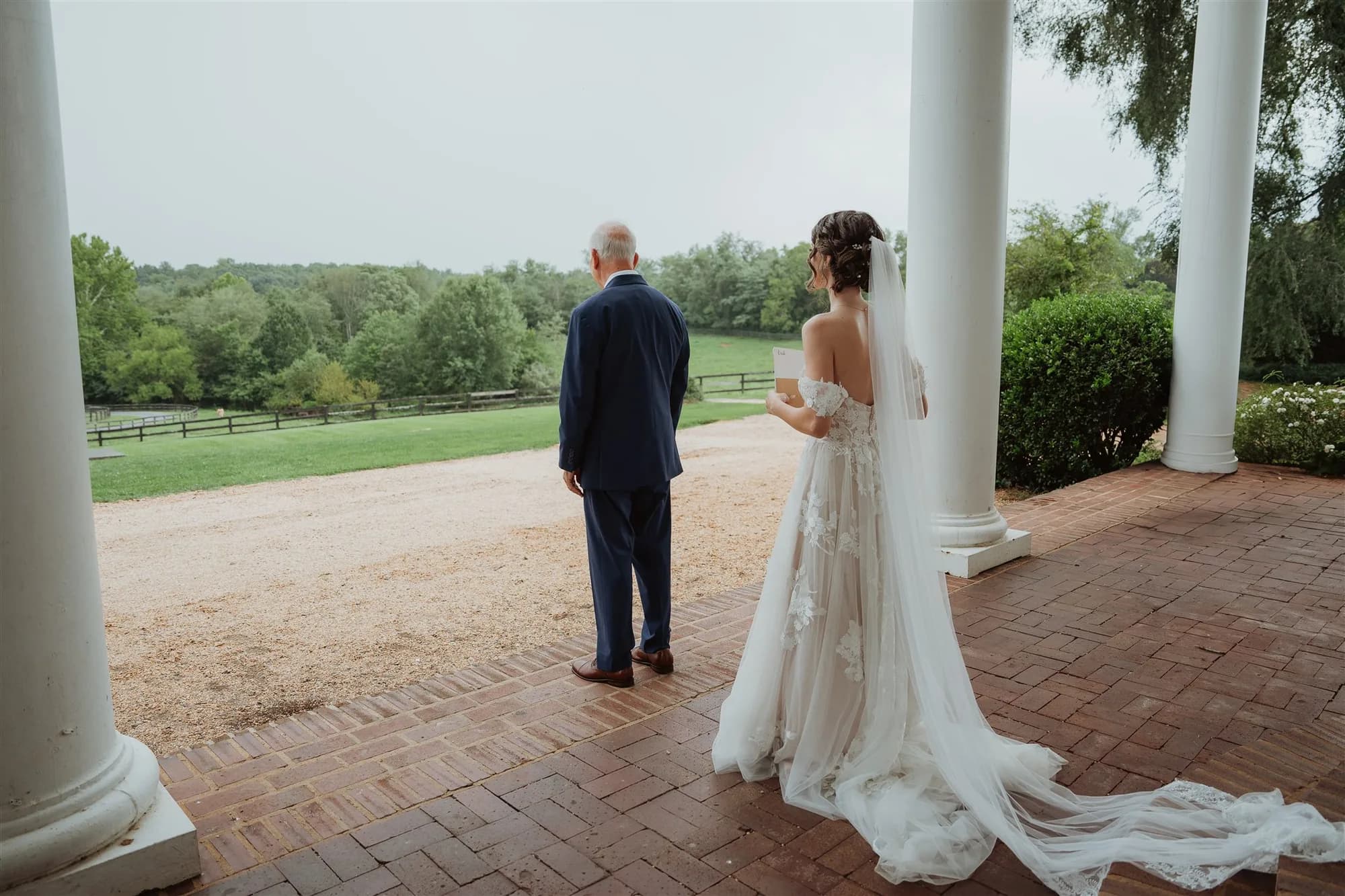 Bride and groom in formal western attire standing on brick patio between white columns overlooking green grounds
