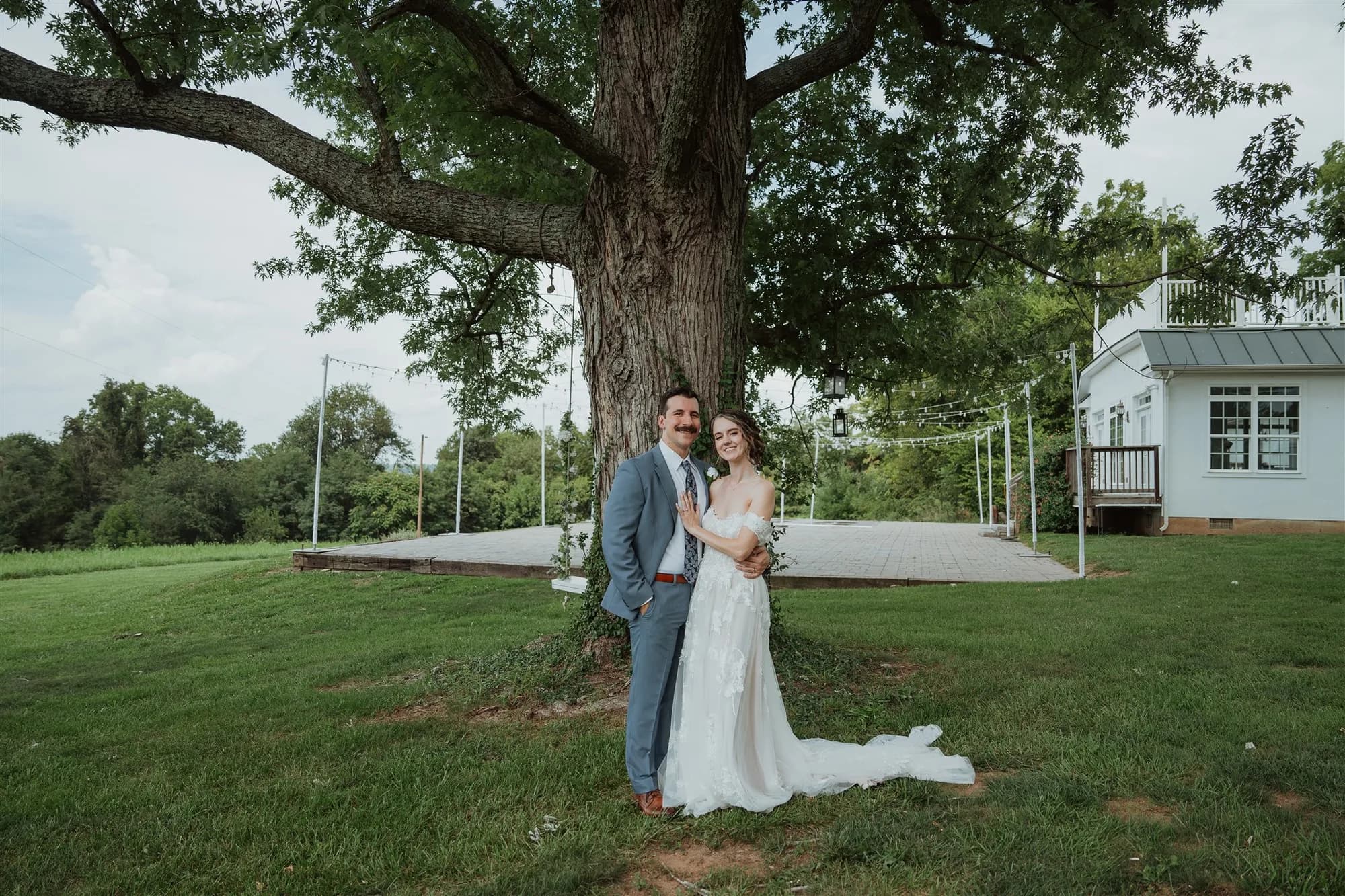 Bride and groom embrace beneath a grand oak tree on Rixey Manor's lush grounds with string lights and white venue building