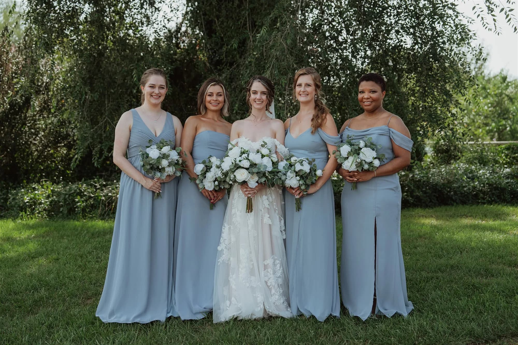 Bride with four bridesmaids in blue dresses holding white flower bouquets outdoors