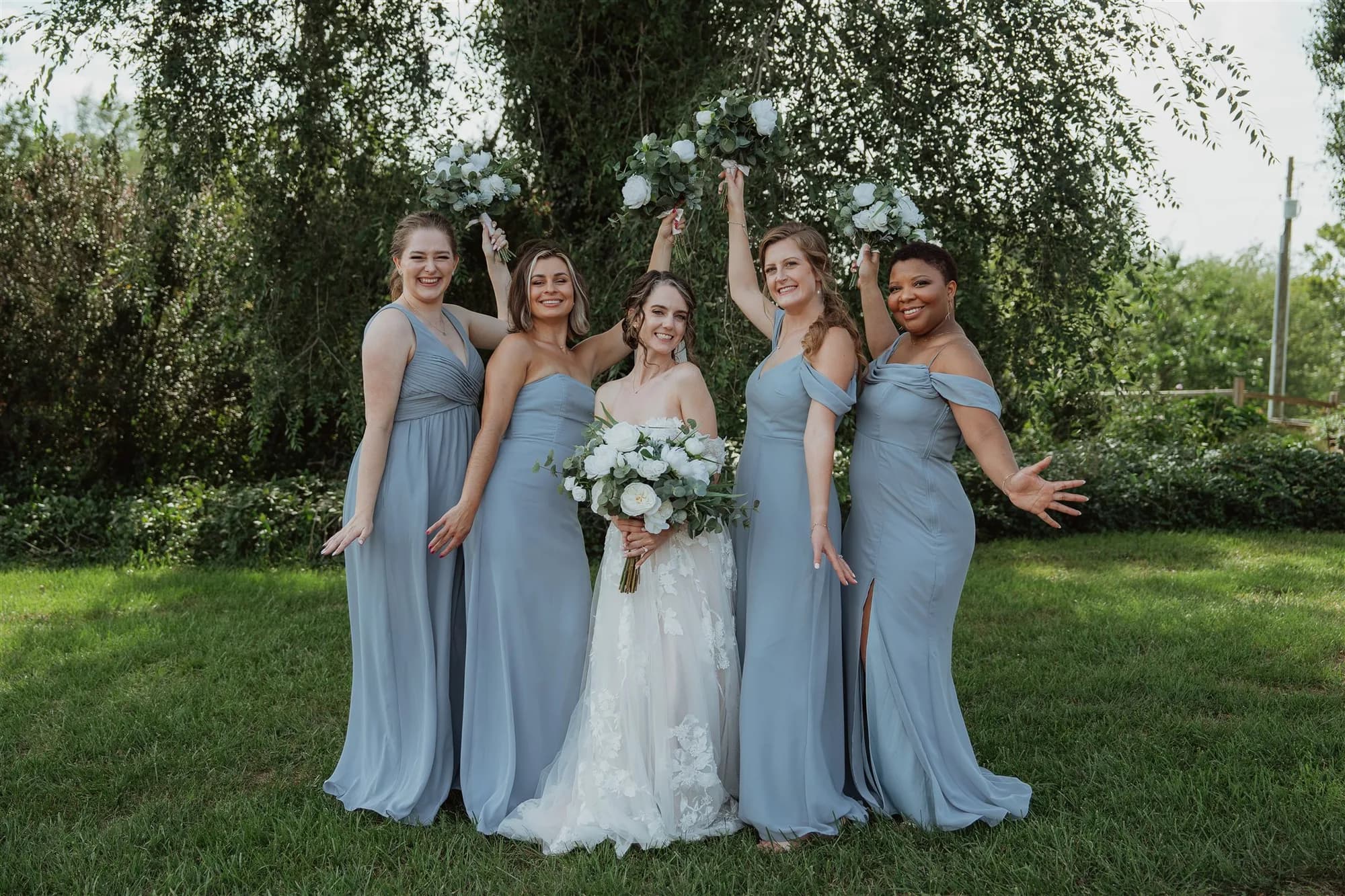 Bride and four bridesmaids in dusty blue gowns laughing outdoors under lush green trees