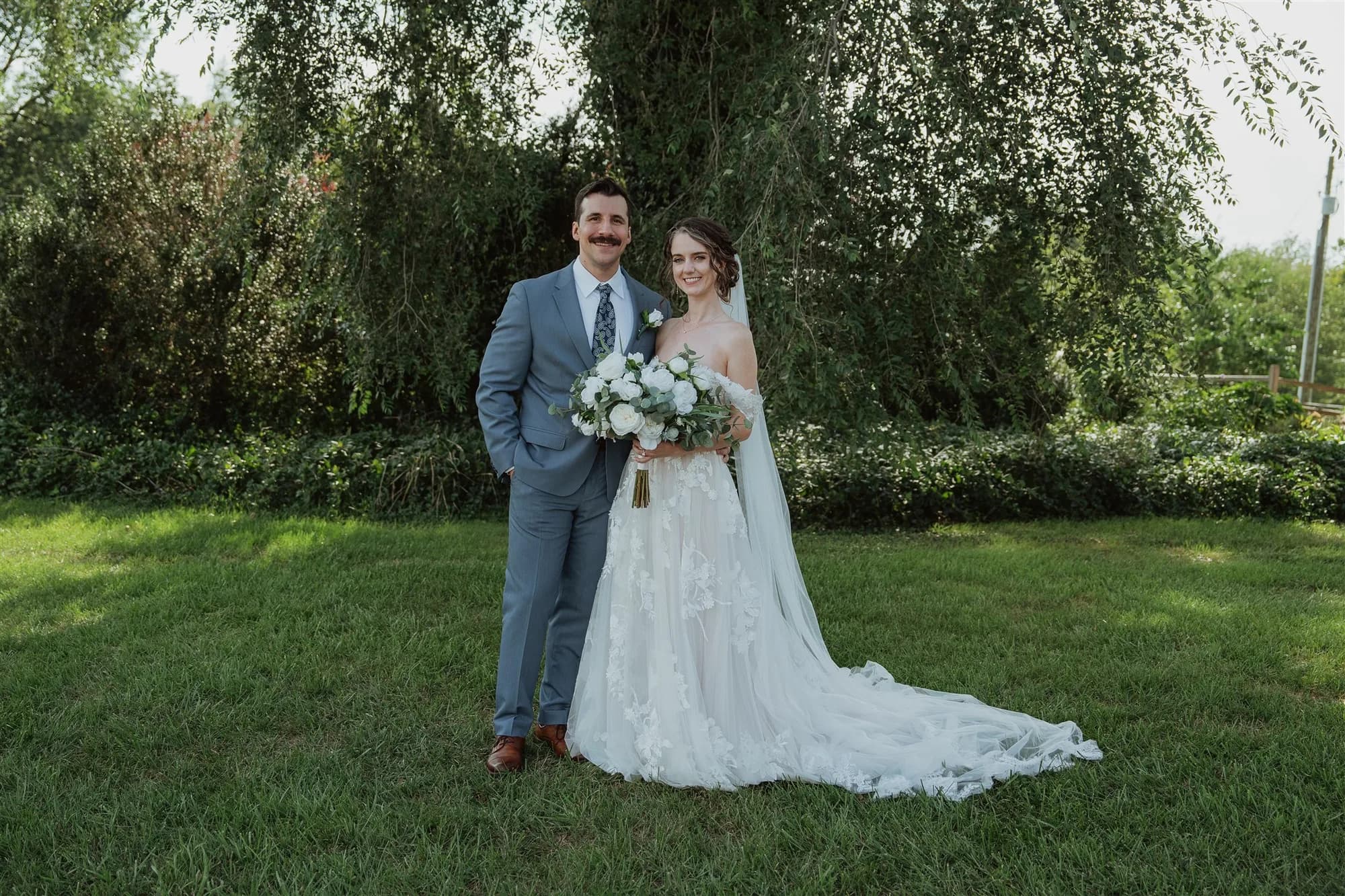 Bride and groom portrait on green lawn beneath a weeping willow, bride holding white bouquet