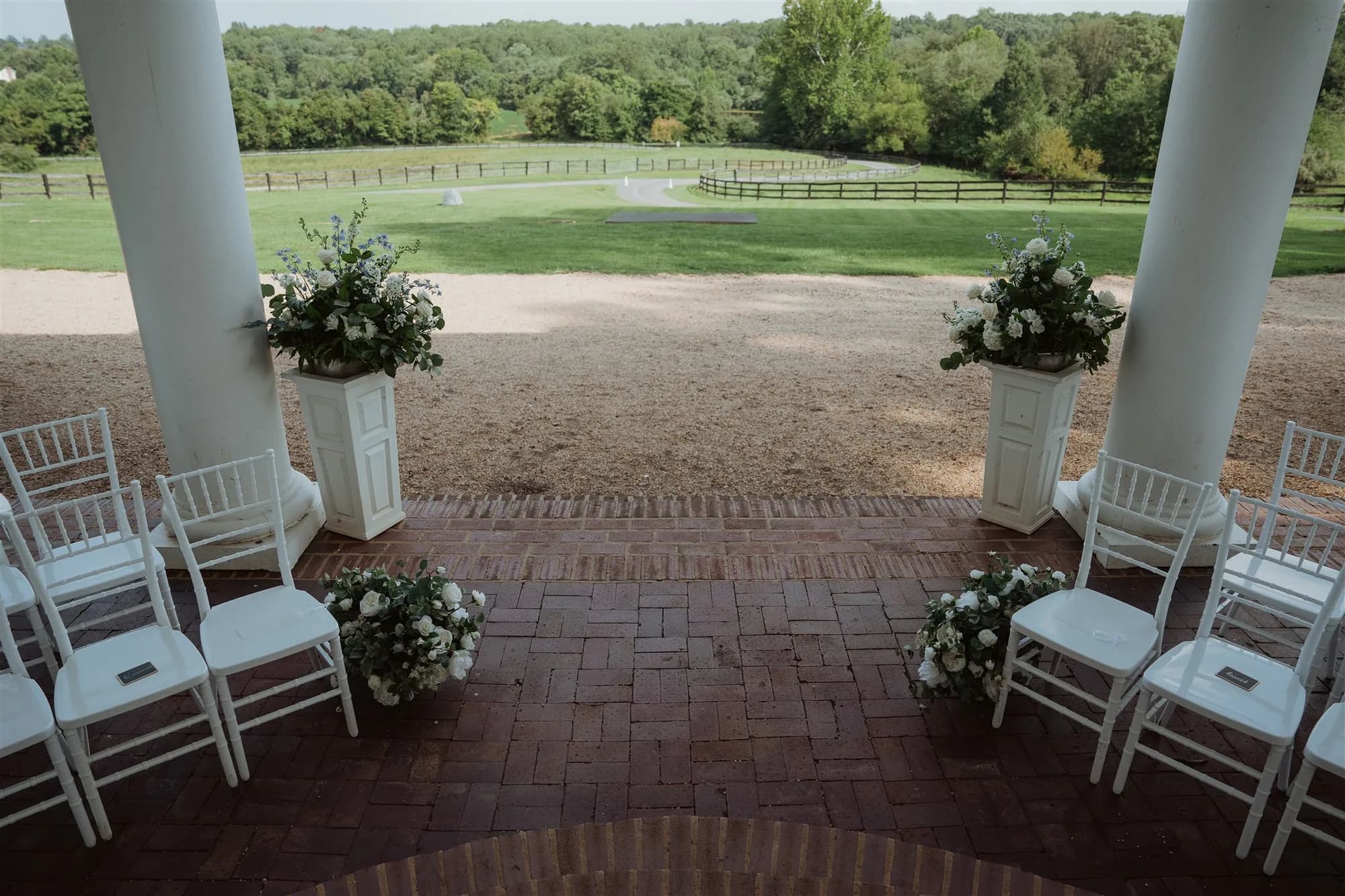 White chiavari chairs and floral pedestals frame a brick ceremony terrace overlooking Rixey Manor's pastoral Virginia grounds