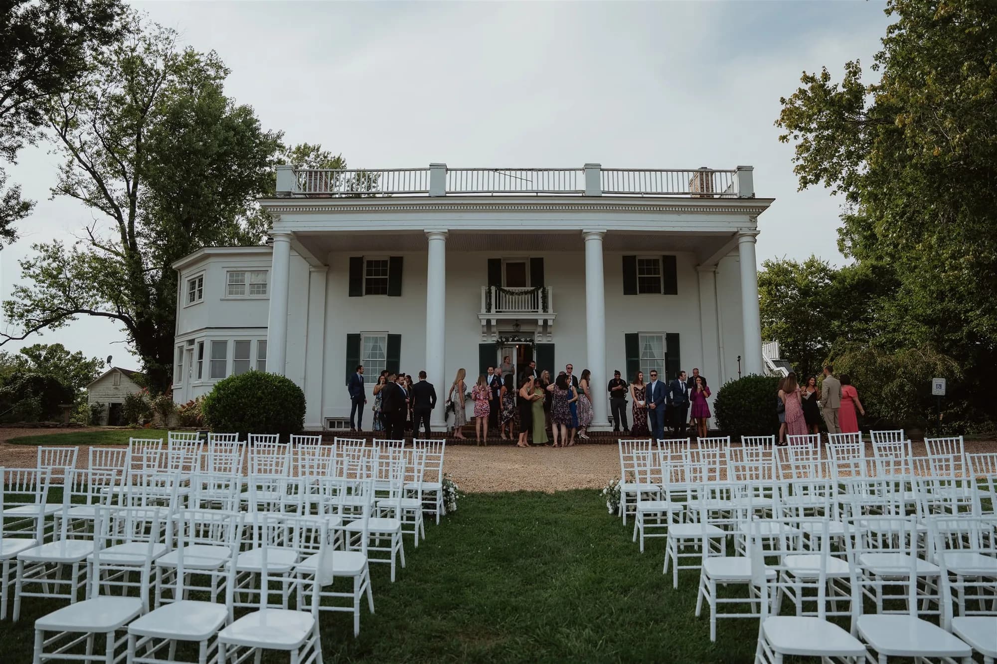 White chiavari chairs arranged on lawn before Rixey Manor's columned facade with wedding guests gathering at entrance