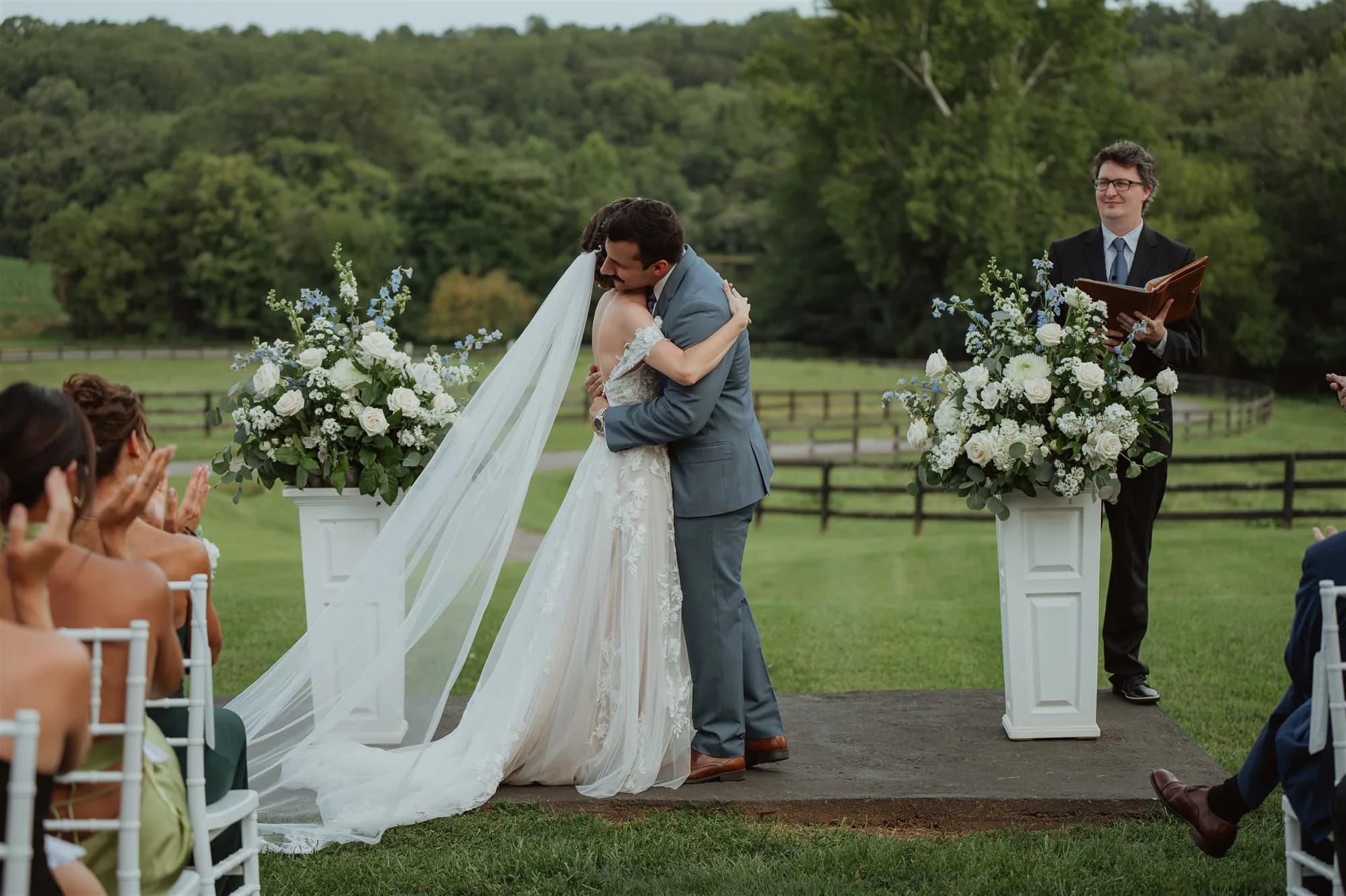 Bride and groom share first kiss embrace at outdoor Rixey Manor ceremony amid white floral arrangements and rolling Virginia hills