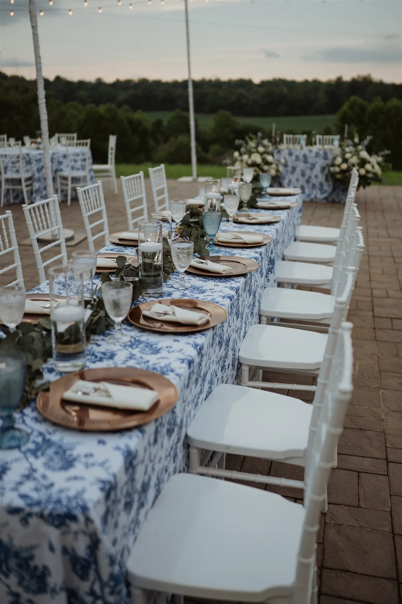 Elegant outdoor reception setup with blue and white patterned table runners, place settings, and white chiavari chairs along long tables.