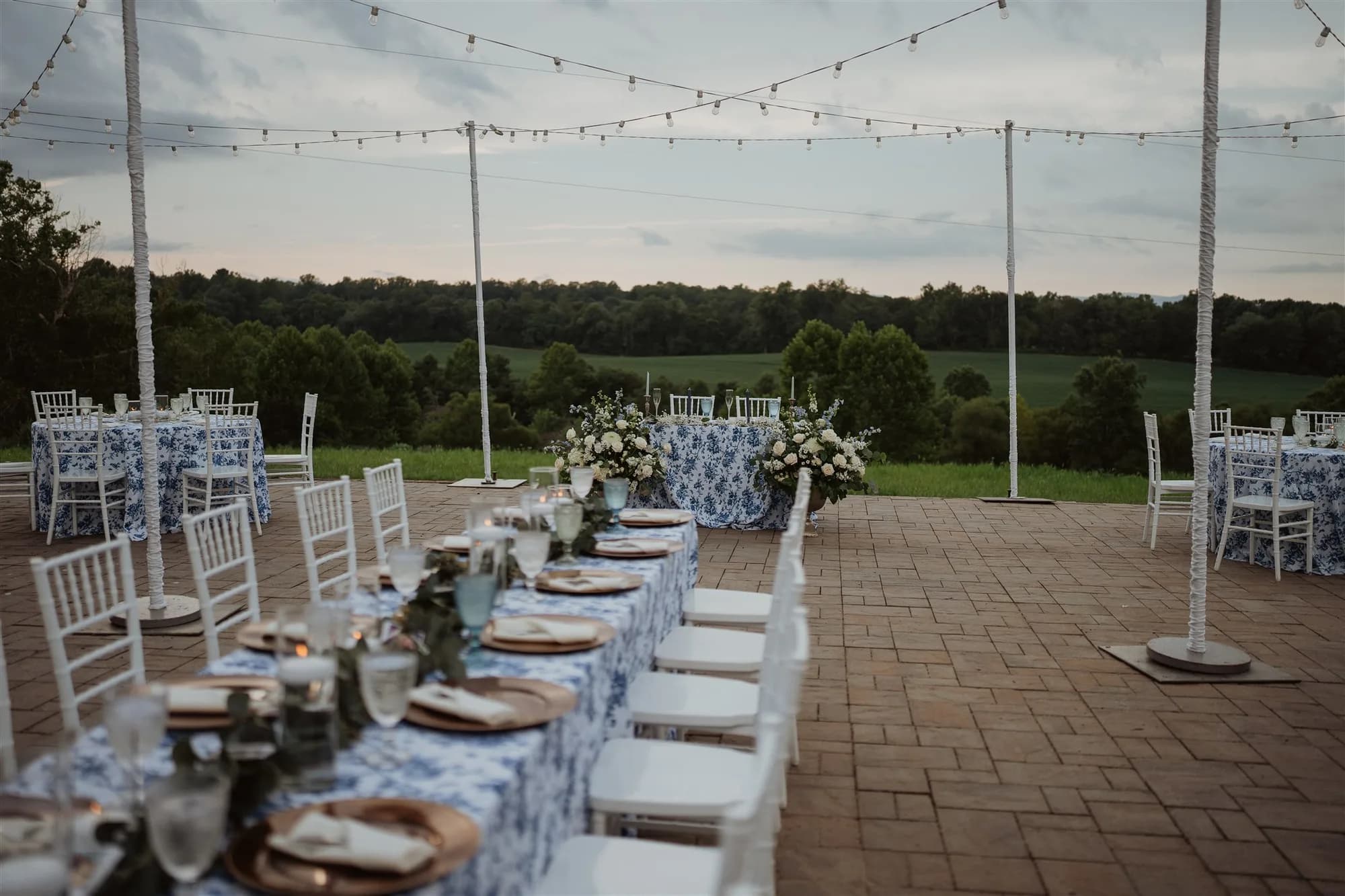 Elegant outdoor reception tables with blue floral linens and string lights overlooking green Virginia countryside at dusk