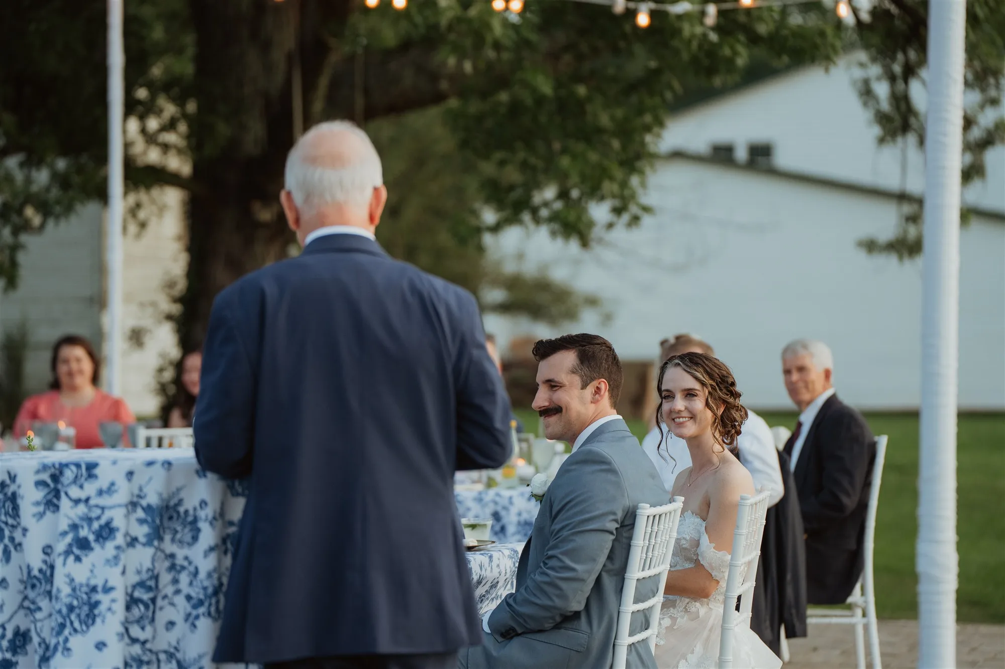 Bride smiles warmly at speaker during outdoor wedding toast as groom looks on at candlelit reception table