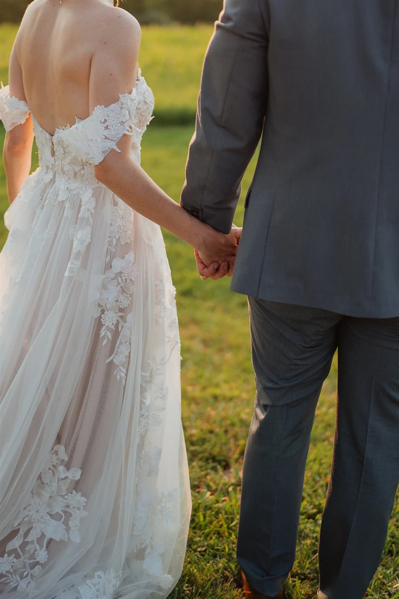 Bride and groom hold hands from behind on green grounds, bride in floral lace gown at golden hour