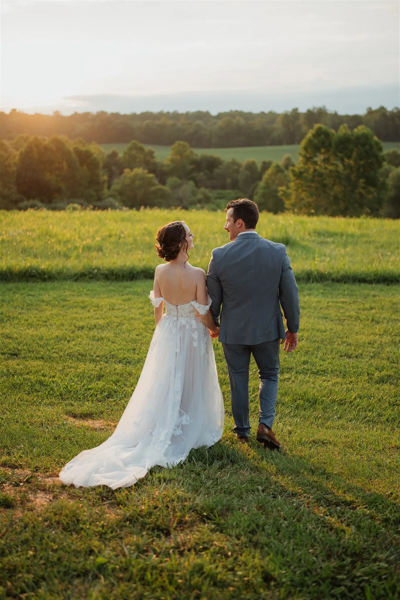 Bride and groom stand together in a green field at golden hour, overlooking rolling countryside.