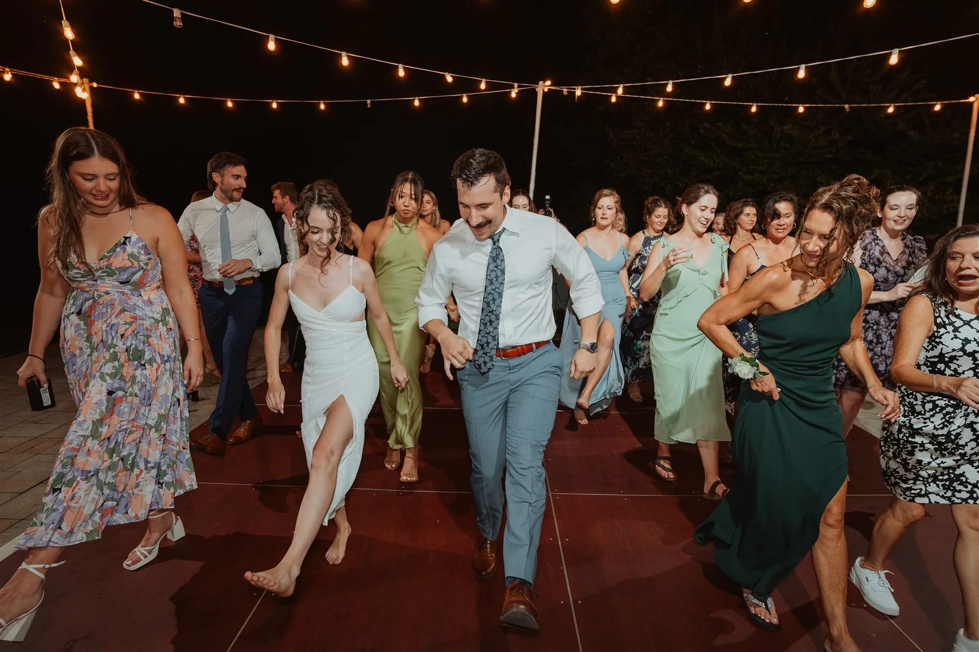 Bride and groom dance energetically with guests under string lights at outdoor wedding reception