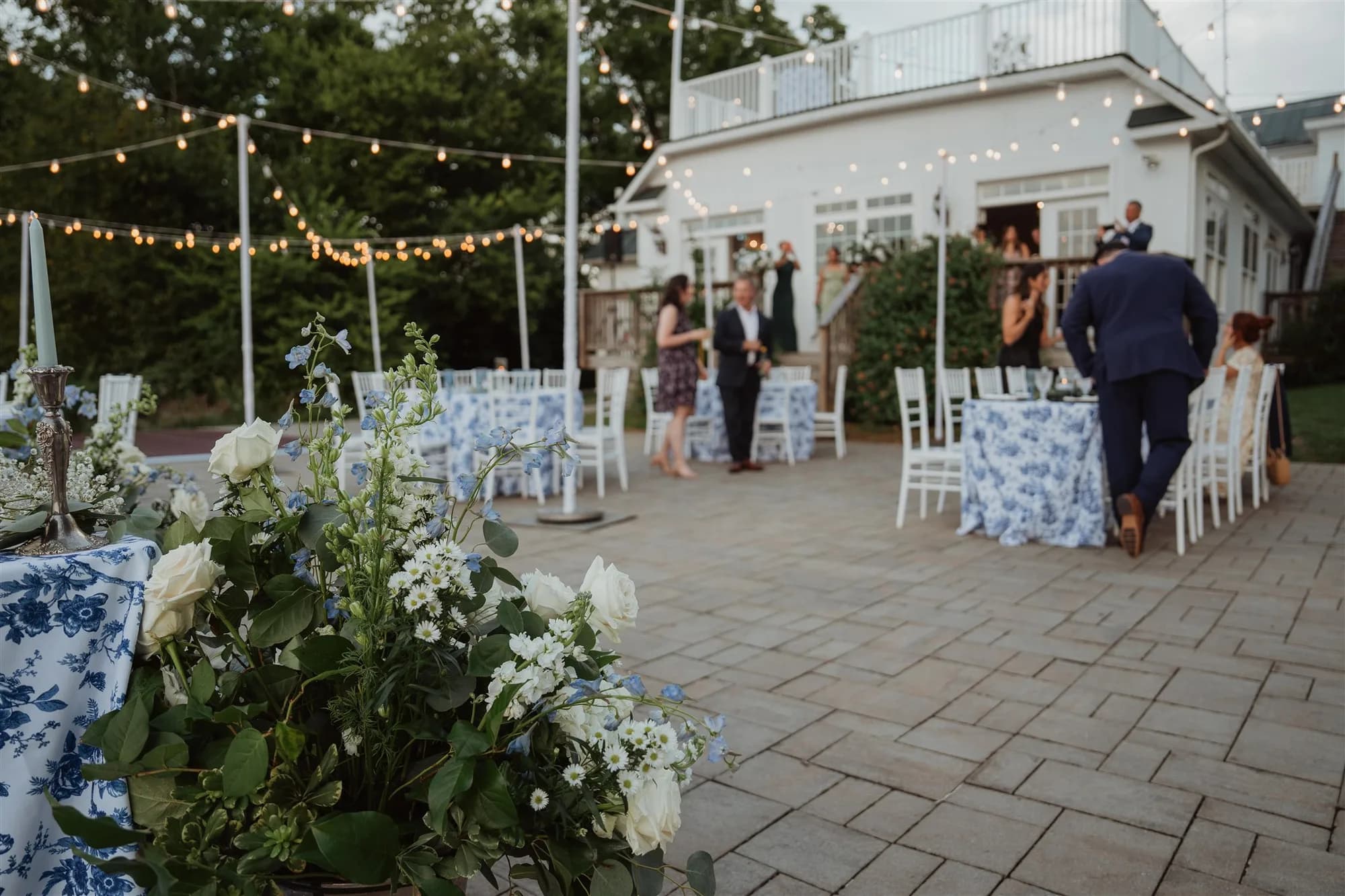 Guests mingle on the Rixey Manor terrace at dusk with string lights, blue floral linens, and white roses in foreground.