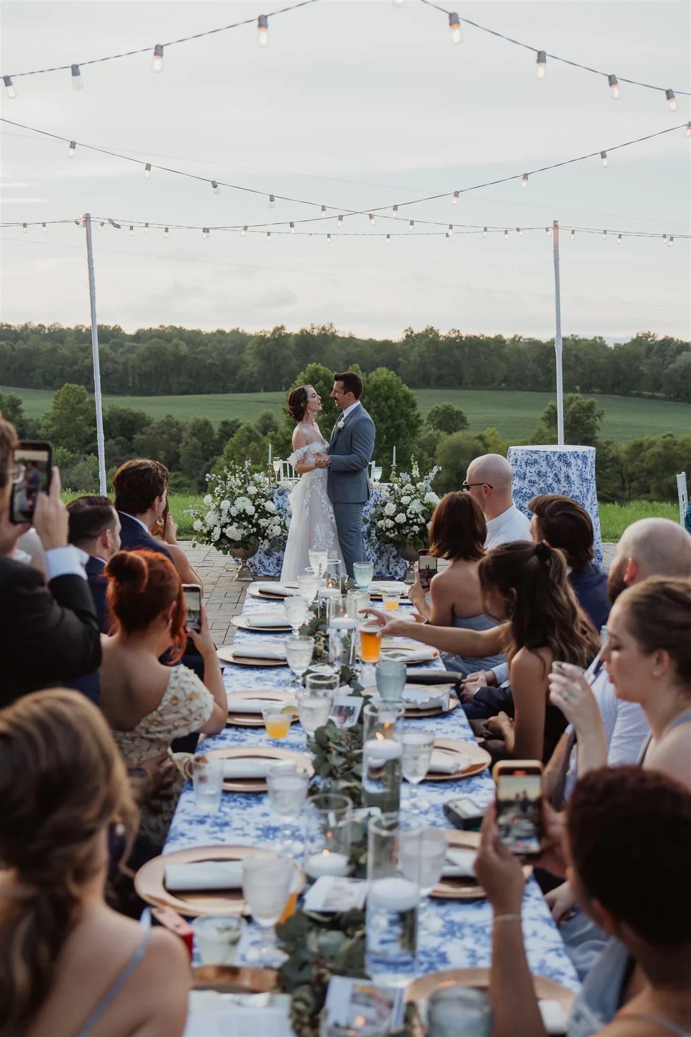 Bride and groom share a moment at the head of a long reception table under string lights with rolling Virginia hills behind them.