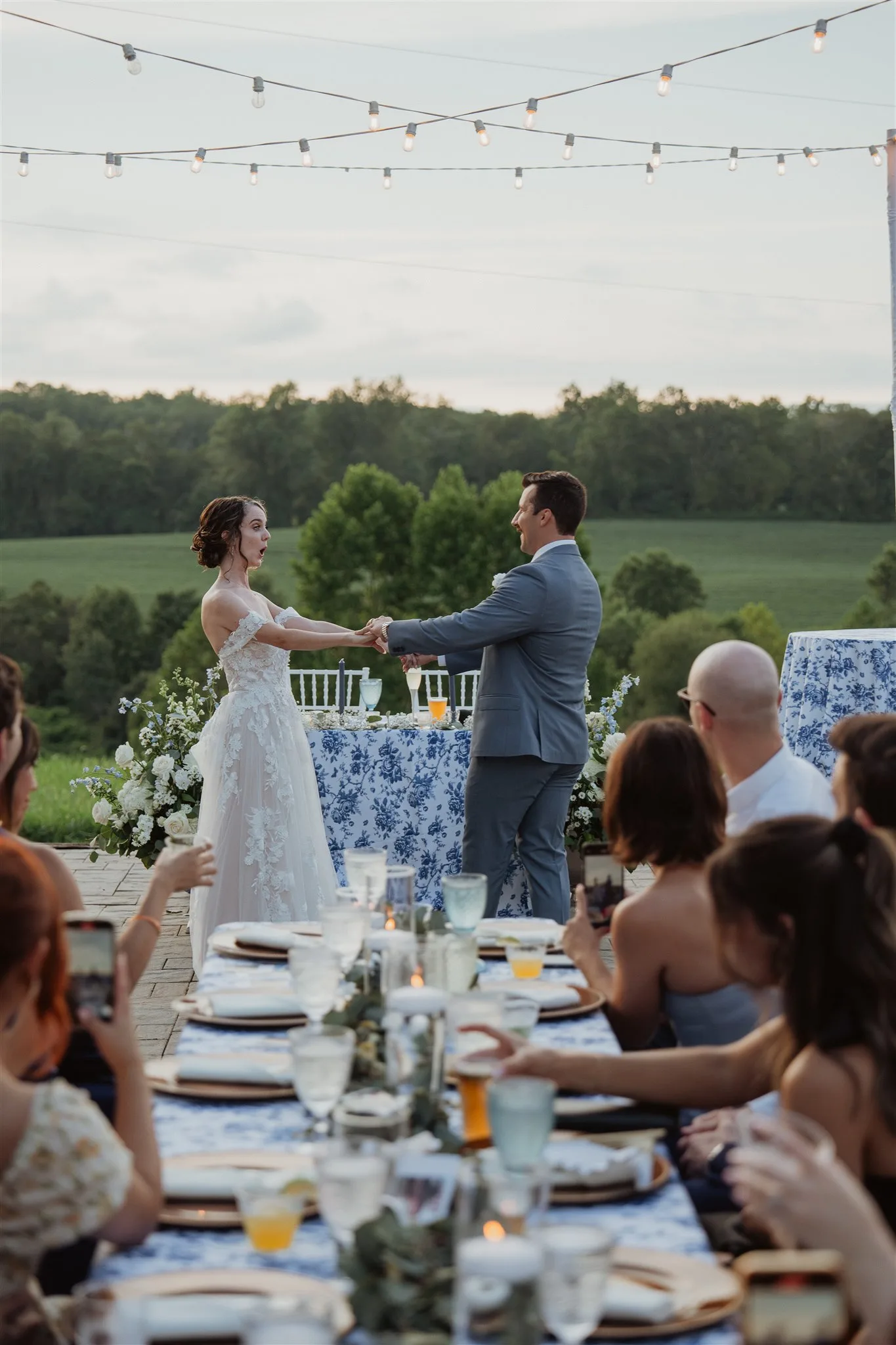 Bride and groom share first dance on outdoor terrace at Rixey Manor, guests at floral table, string lights and rolling fields behind