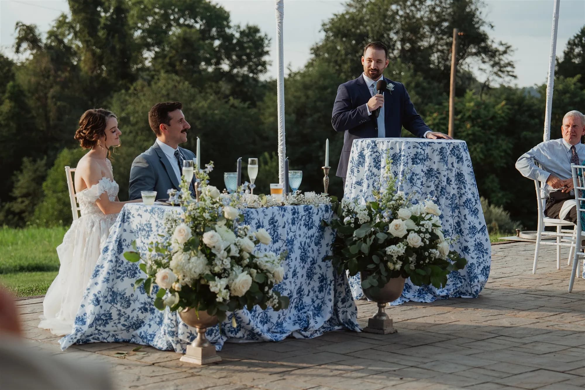 Best man gives toast as bride and groom listen at outdoor reception table with blue floral linens at Rixey Manor