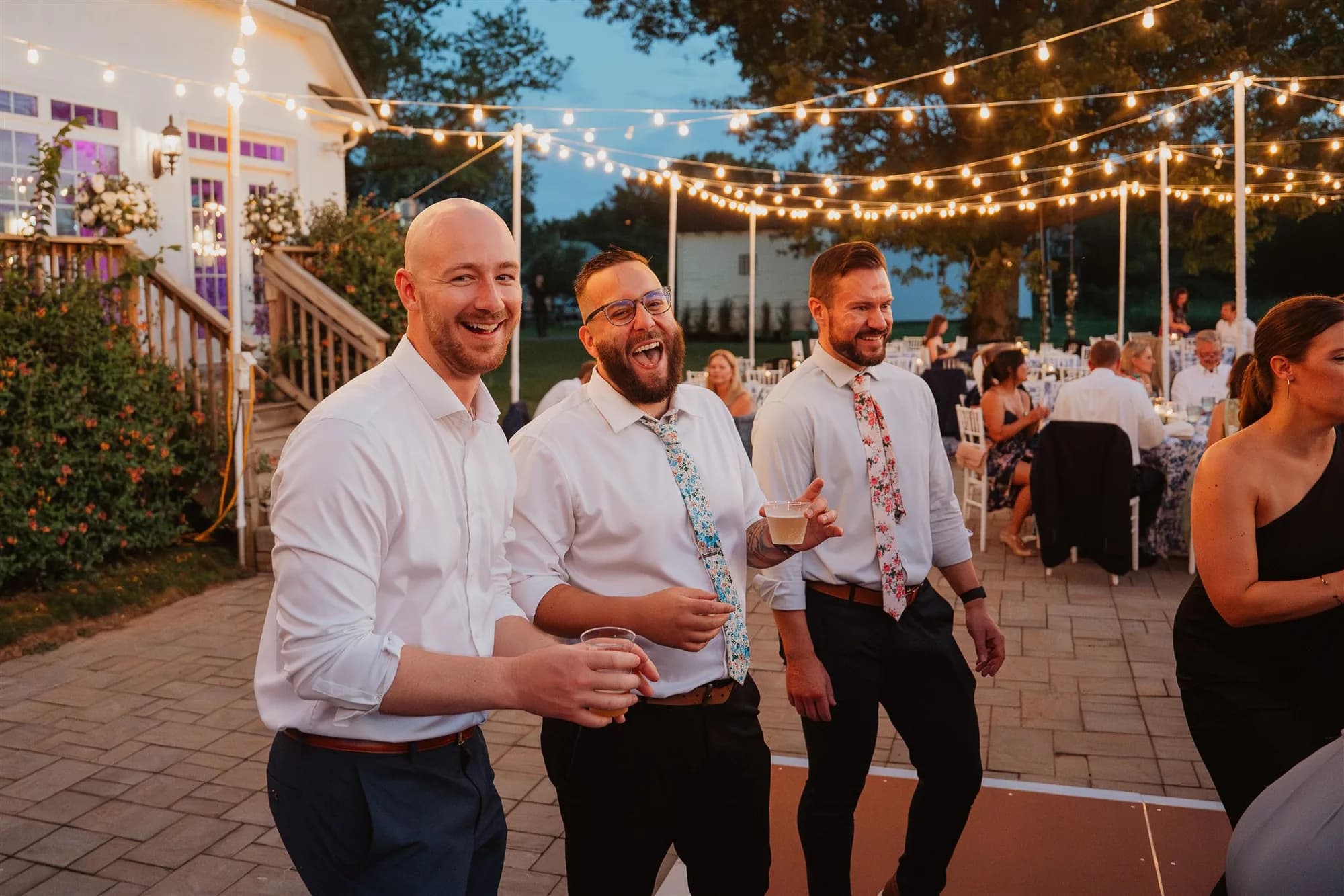 Three laughing groomsmen on the Rixey Manor terrace under string lights at dusk