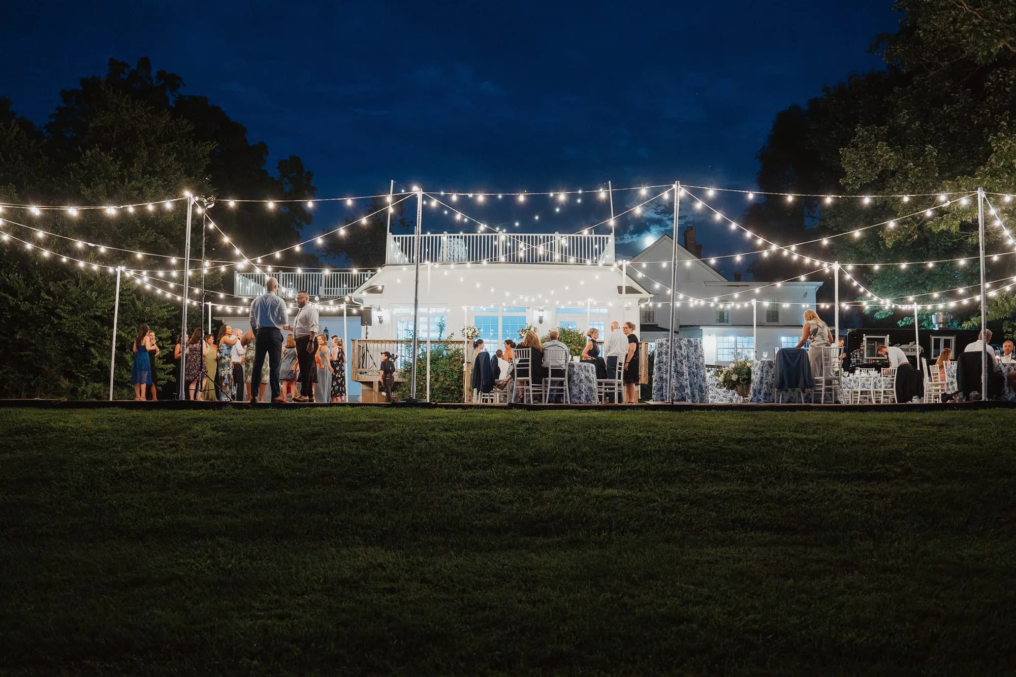 Evening wedding reception under string lights with white manor building illuminated in background, guests gathered on lawn
