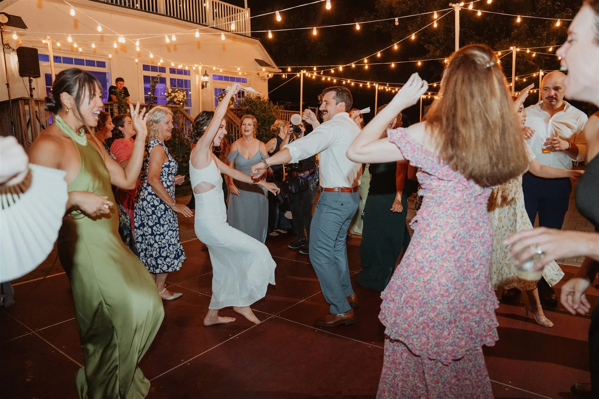 Bride and groom dance joyfully with guests under string lights on an outdoor patio at night