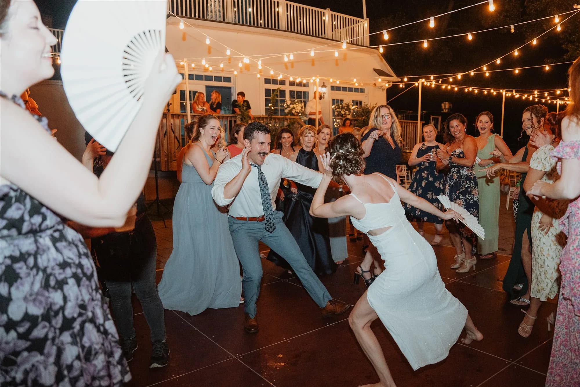 Bride and groom dance dramatically on outdoor reception patio as excited guests cheer under string lights at night.