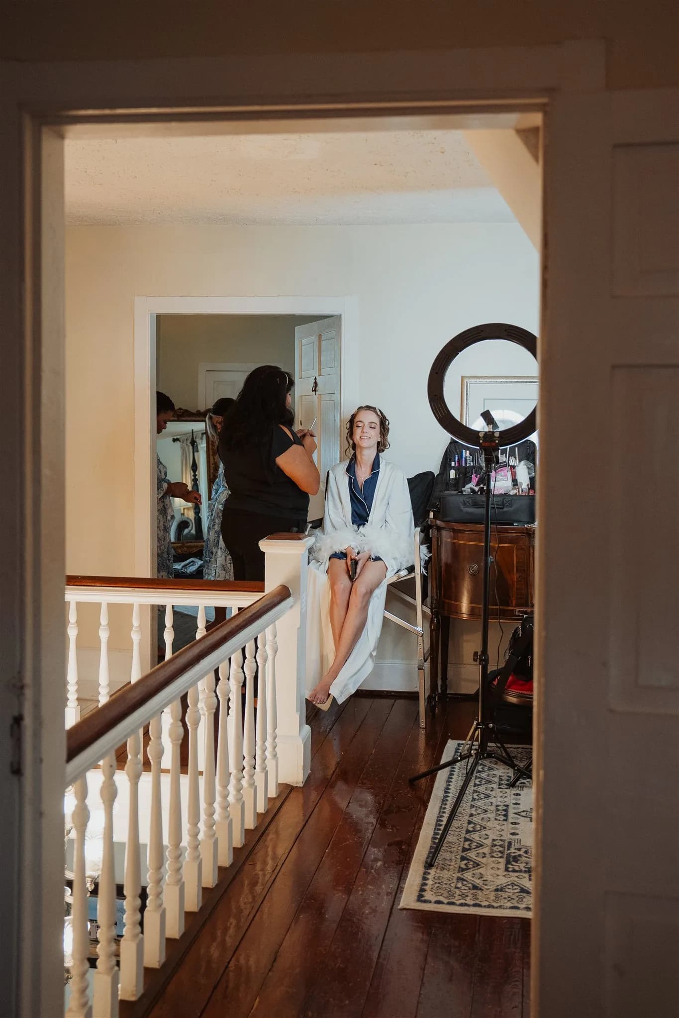 Bride in white robe getting ready in manor hallway with ring light and mirror setup