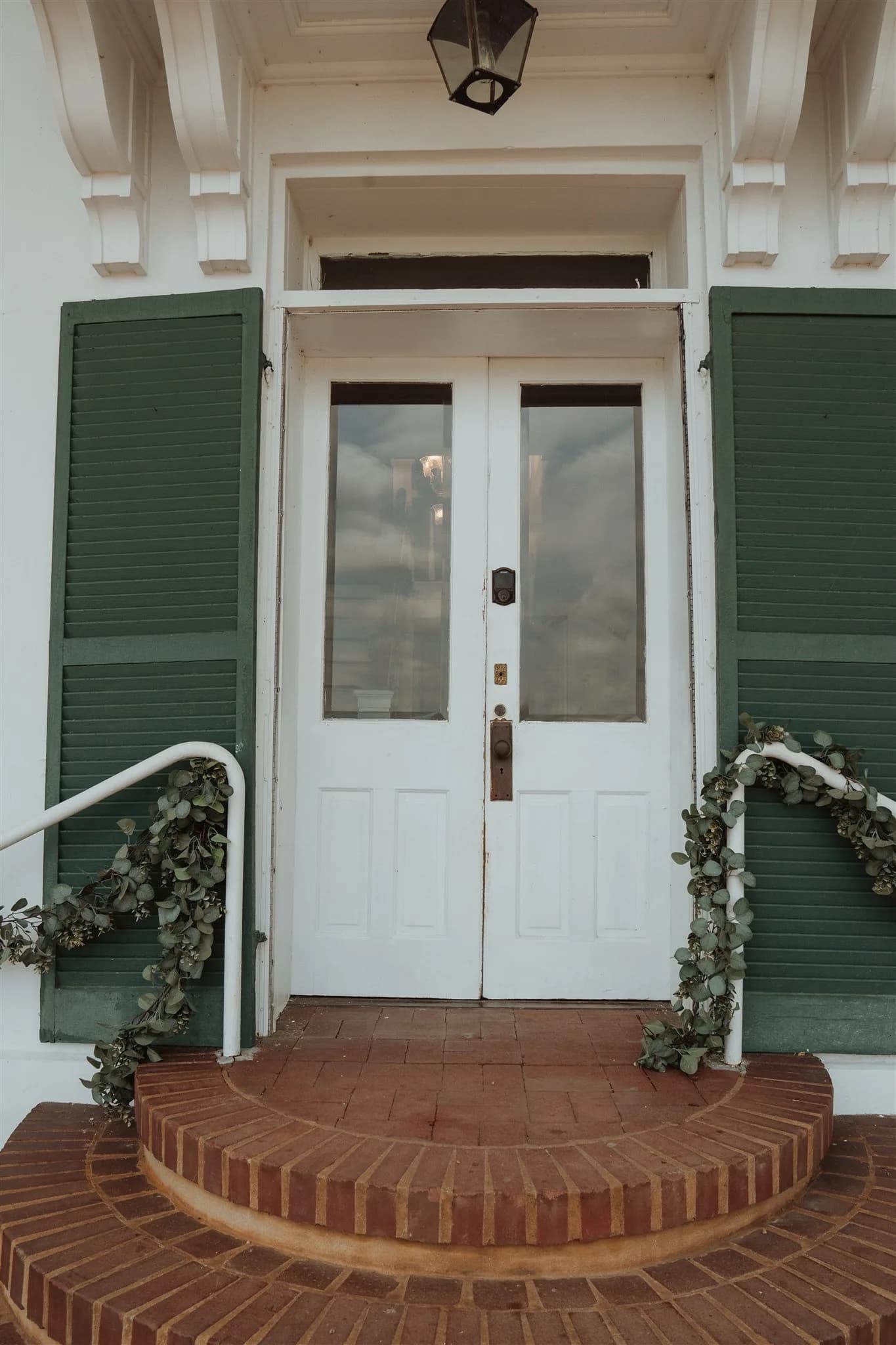 White double doors with green shutters and eucalyptus garland on brick steps at Rixey Manor entrance